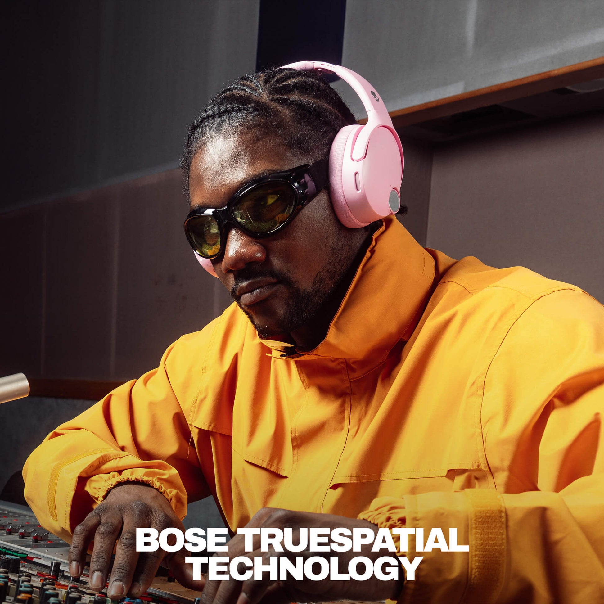 Person wearing pink Skullcandy Crusher 1080 ANC headphones and sunglasses, sitting at a mixing console with 'Bose TrueSpacial Technology' text. | Candy
