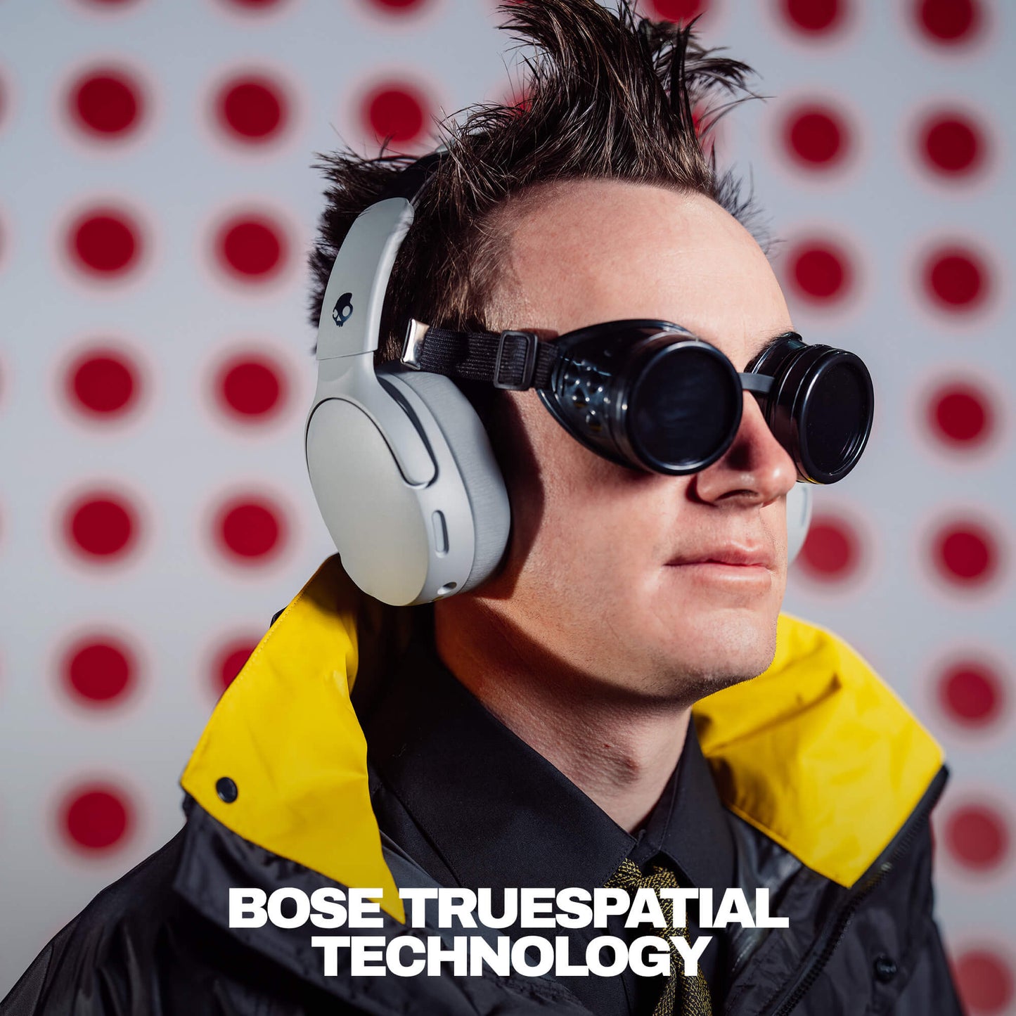 Person wearing Grey Skullcandy Crusher 1080 ANC headphones and goggles with 'Bose TrueSpacial Technology' text against a red and white polka dot background. | Cement