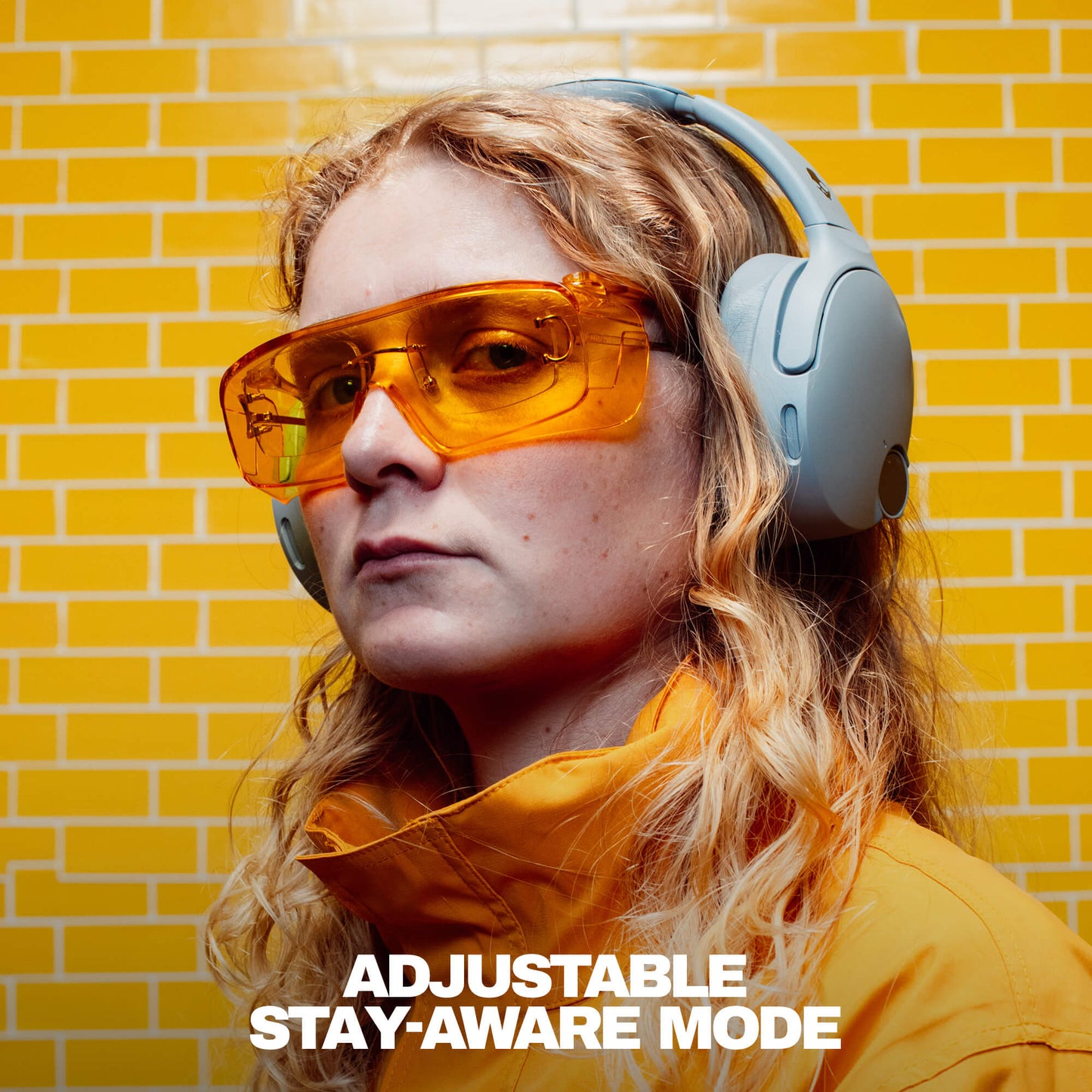 Person wearing Skullcandy Crusher 1080 ANC Headphones, orange sunglasses and a yellow jacket with a yellow brick wall background | Cement