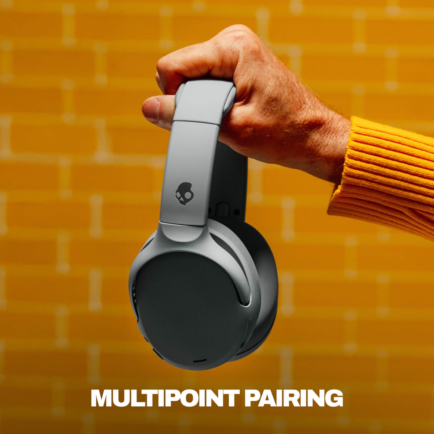 Hand holding a pair of headphones with 'MultiPoint Pairing' text on a yellow brick wall background | Cement