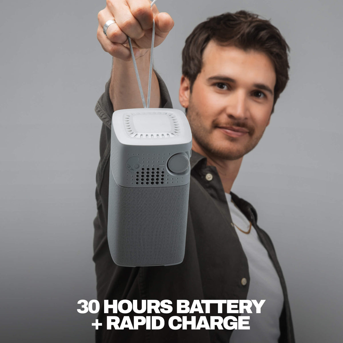 A man holding up a gray session 900 with a hanging strap against a gray studio background, with text at the bottom reading '30 HOURS BATTERY + RAPID CHARGE'. | Cement