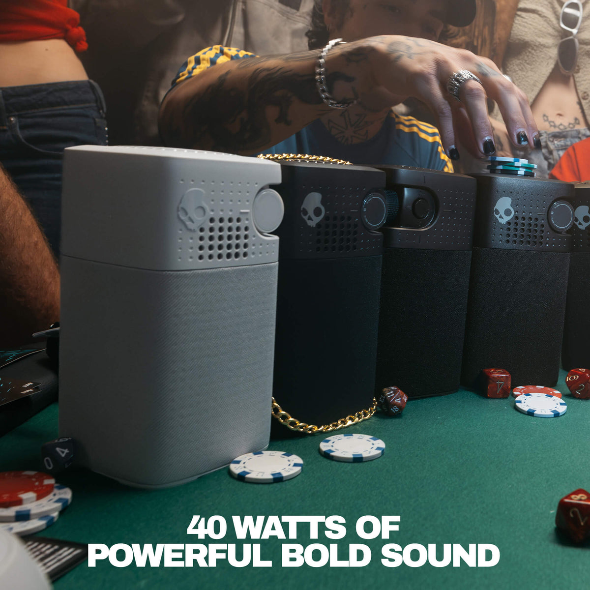 Three Skullcandy portable speakers in gray and black are lined up on a green felt table with poker chips and dice, with a person's tattooed hand reaching toward the devices. Text at the bottom reads '40 WATTS OF POWERFUL BOLD SOUND'. | Cement