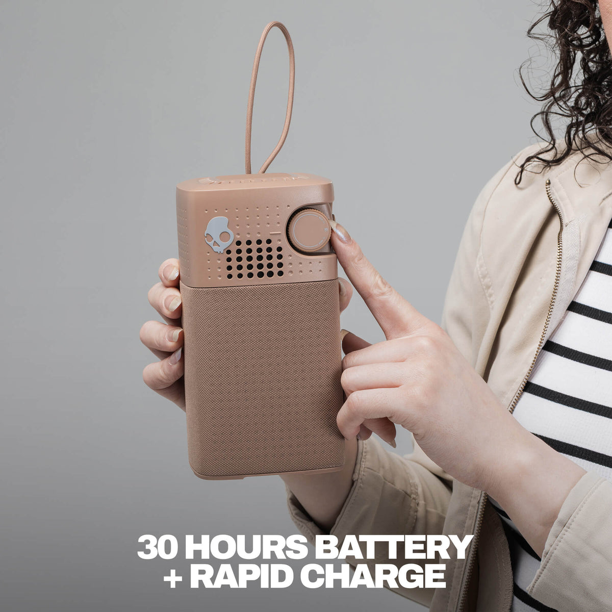 A person holds a session 900 speaker in a warm beige/tan color against a plain gray background. The person is wearing a beige jacket over a black-and-white striped shirt. | Primer