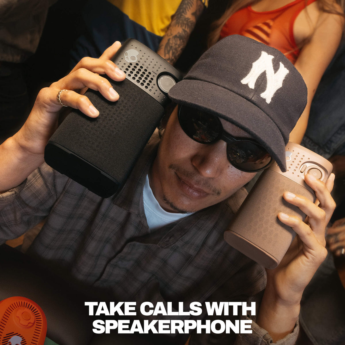 A man wearing a black baseball cap with a white 'N' logo, sunglasses, and a plaid shirt holds two session 900 speakerphones, one black and one taupe, to his ears. The text 'TAKE CALLS WITH SPEAKERPHONE' is displayed at the bottom. | True Black