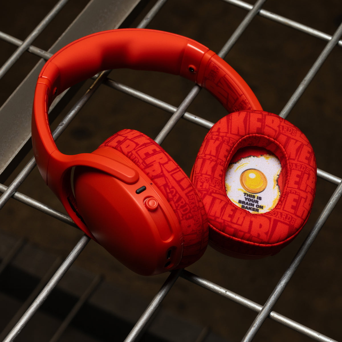 Red over-ear headphones with textured ear cushions and a novelty egg graphic on the inner right earcup, resting on a metal grid surface. | Skullcandy X Baker Skateboards