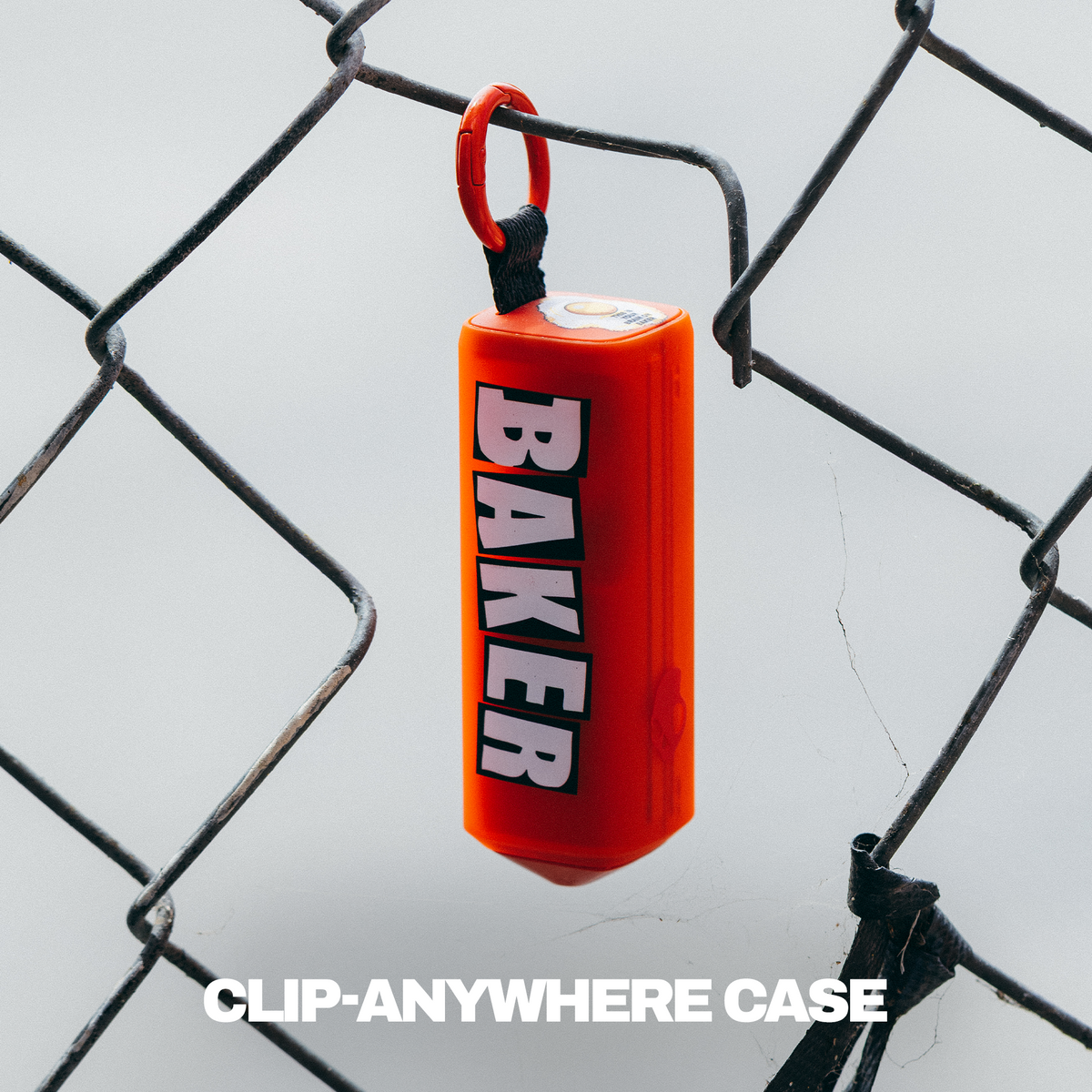A bright orange cylindricalDime 360 case with the word 'BAKER' printed vertically in white and black, hanging from a black loop attached to a chain-link fence. The text 'CLIP-ANYWHERE CASE' appears at the bottom. | Skullcandy X Baker Skateboards