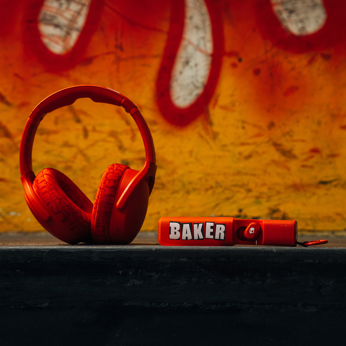 Red over-ear Crusher 540 headphones and a red dime 360 earbuds with the word 'BAKER' in white letters, placed on a dark ledge against a graffiti-covered orange and yellow wall. | Skullcandy X Baker Skateboards