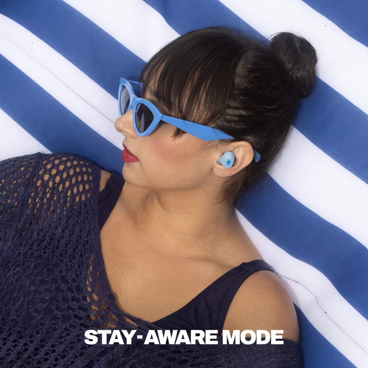 A woman wearing light blue cat-eye sunglasses and matching light blue Dime 360 earbuds, lying on a blue and white striped cushion. The text 'STAY-AWARE MODE' is displayed at the bottom. | Skullcandy X Peanuts Lucy