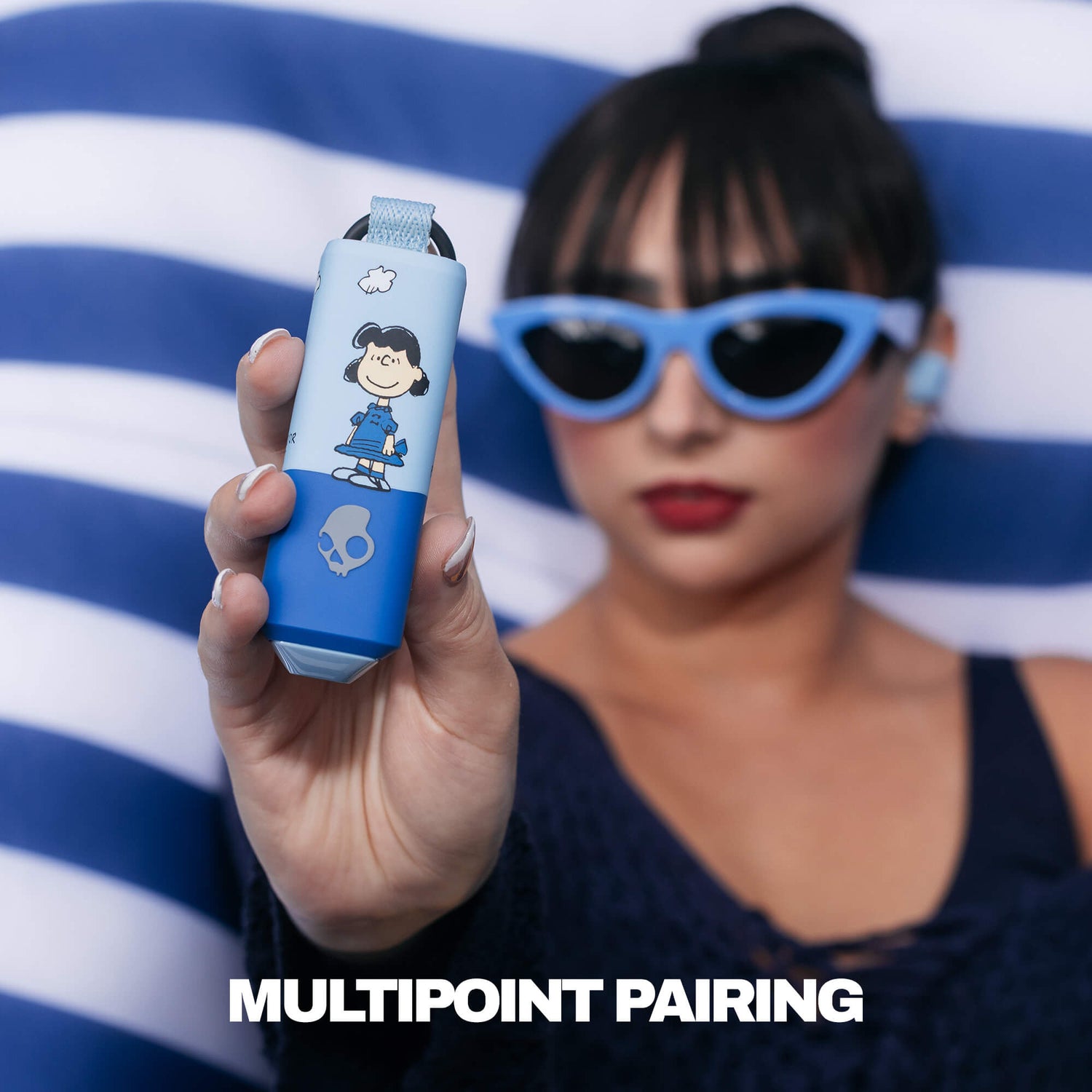 A woman in blue cat-eye sunglasses holds up a light blue and blue-gray Dime 360 earbuds featuring Lucy from peanuts and a skull graphic. The background is a blue and white striped pattern, and the text 'MULTIPONT PAIRING' is displayed at the bottom. | Skullcandy X Peanuts Lucy