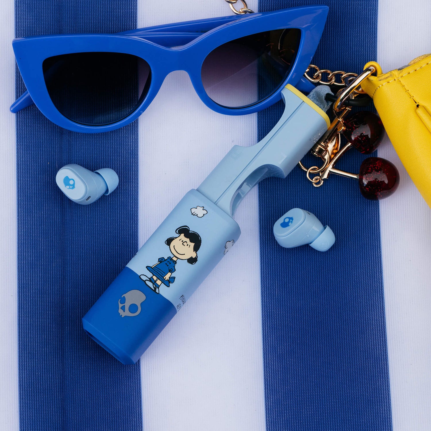 A styled flat-lay featuring a light blue and blue dime 360 earbuds with Lucy from peanuts on it, blue sunglasses, and a yellow purse on a blue and white striped background. | Skullcandy X Peanuts Lucy