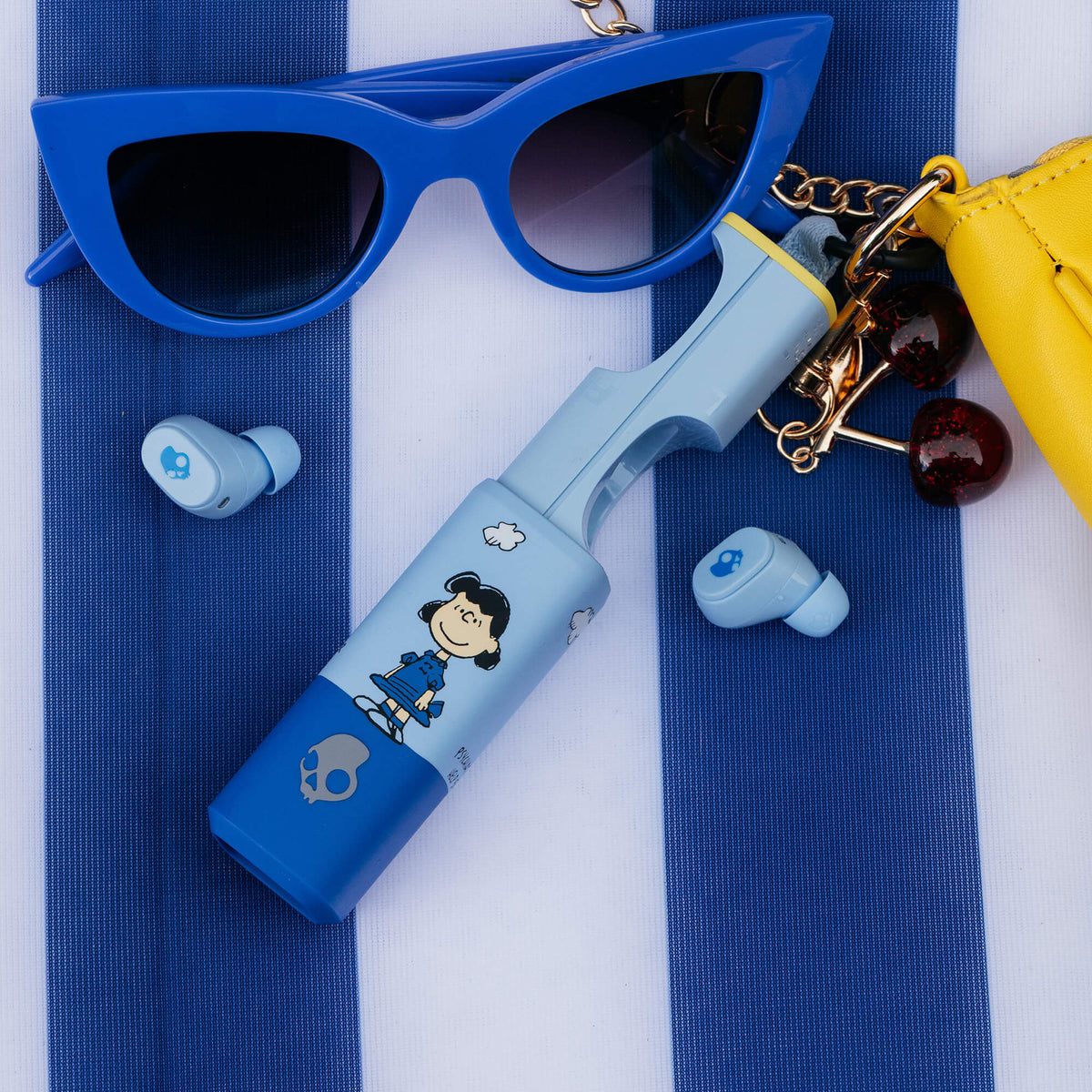 A styled flat-lay featuring a light blue and blue dime 360 earbuds with Lucy from peanuts on it, blue sunglasses, and a yellow purse on a blue and white striped background. | Skullcandy X Peanuts Lucy