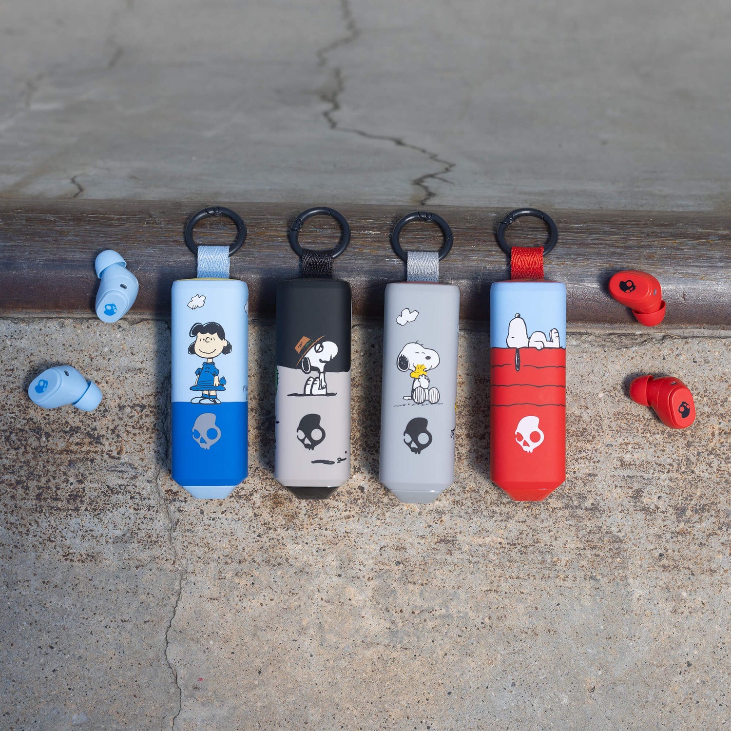 Four Snoopy-themed keychain Dime 360 earbuds in blue, black, gray, and red are lined up on a weathered wooden beam, with matching red and blue earbuds resting on either side on a concrete surface. | Skullcandy X Peanuts Lucy