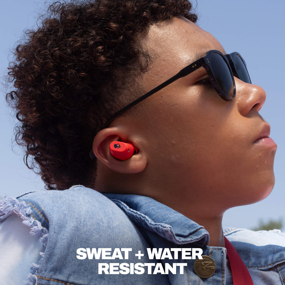 A close-up side profile of a person outdoors wearing black sunglasses and a red in-ear earbud. The person has curly dark hair and is wearing a denim jacket, with the text 'SWEAT + WATER RESISTANT' overlaid at the bottom. | Skullcandy X Peanuts Snoopy