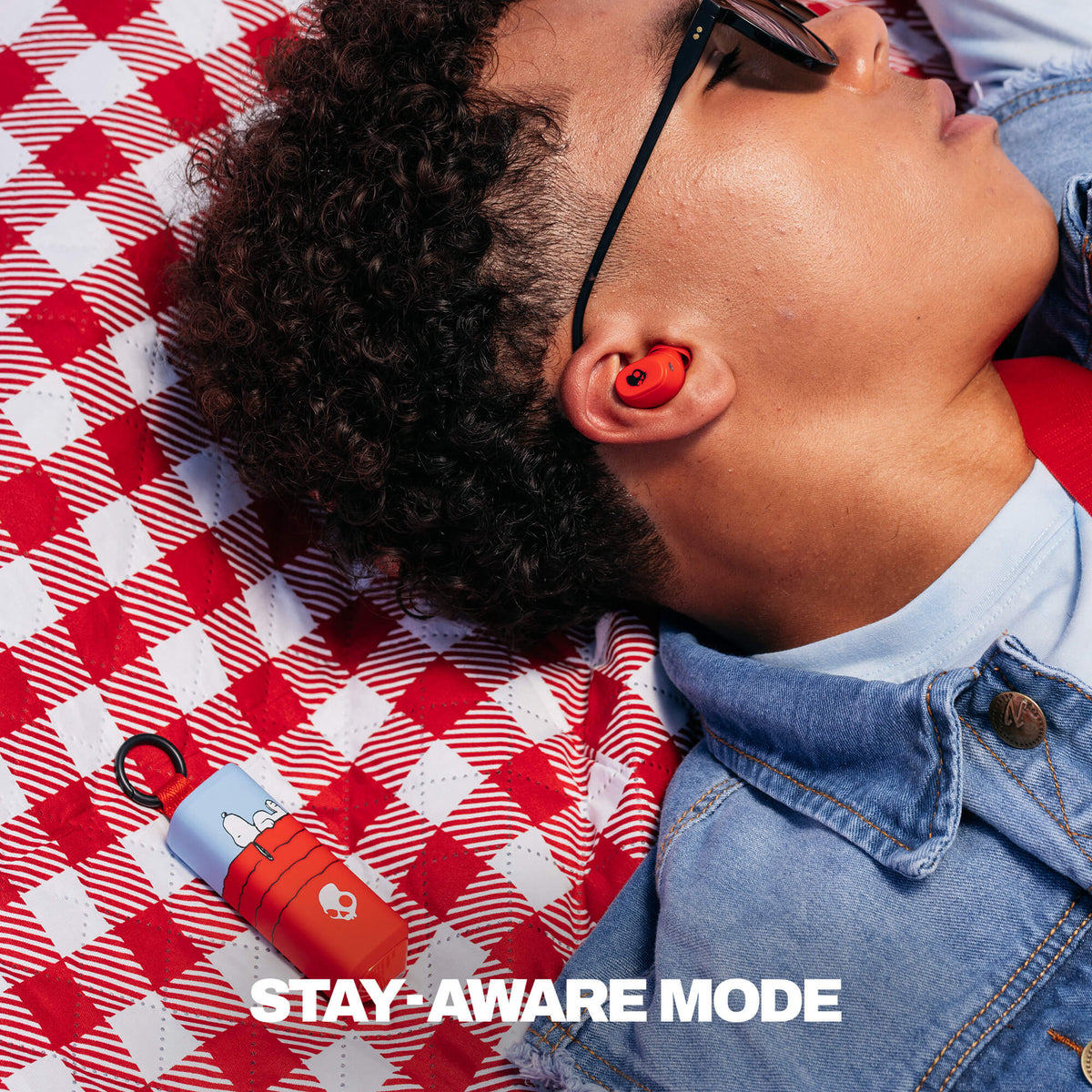 A close-up lifestyle shot of a person lying on a red and white checkered blanket, wearing a red in-ear earbud. A matching red and light blue portable earbud case lies nearby on the blanket, with the text 'STAY-AWARE MODE' overlaid at the bottom. | Skullcandy X Peanuts Snoopy