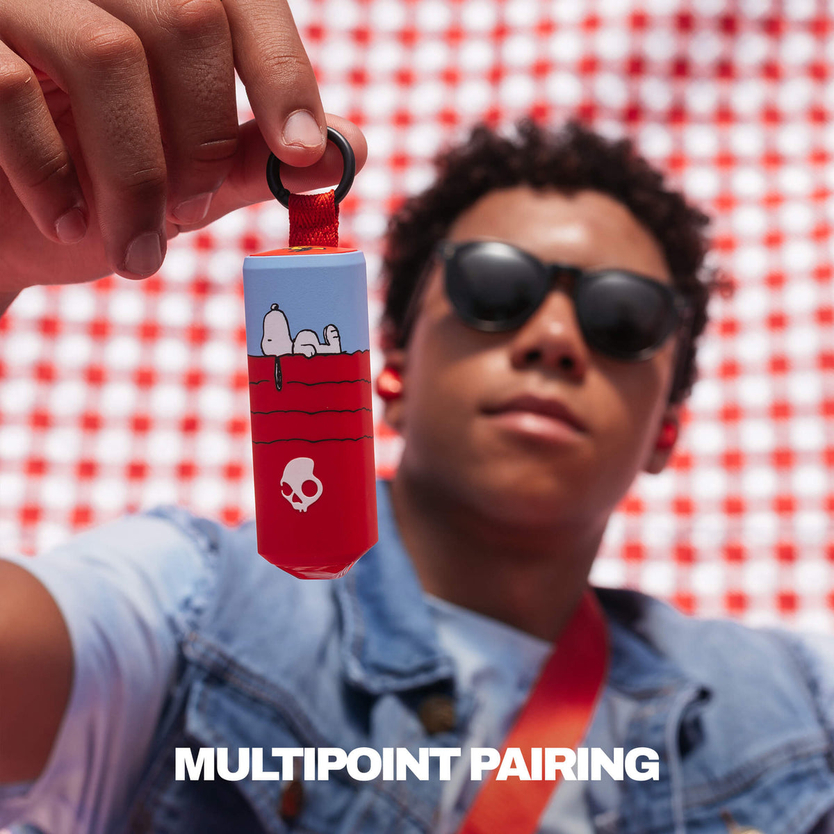 A person in sunglasses and a denim jacket holds up a red and light blue cylindrical Dime 360 or tag with Snoopy and a black ring, against a red-and-white checkered background. The text 'MULTIPONT PAIRING' appears at the bottom. | Skullcandy X Peanuts Snoopy