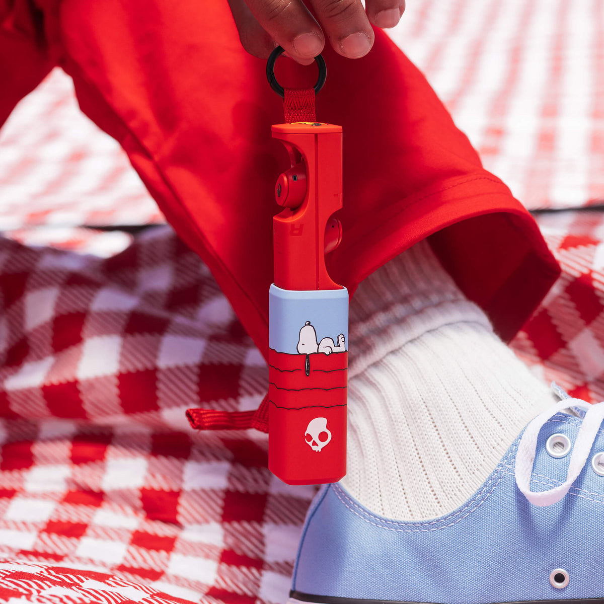 A red and light blue Dime 360, held by a person wearing a red sweatshirt, white sock, and light blue canvas shoe on a red and white checkered blanket. | Skullcandy X Peanuts Snoopy