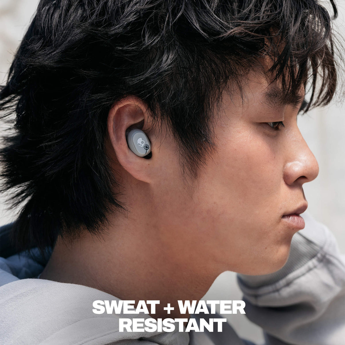 A close-up side profile of a man wearing a gray in-ear wireless earbud, with bold white text at the bottom reading 'SWEAT + WATER RESISTANT'.  | Skullcandy X Peanuts Woodstock & Snoopy