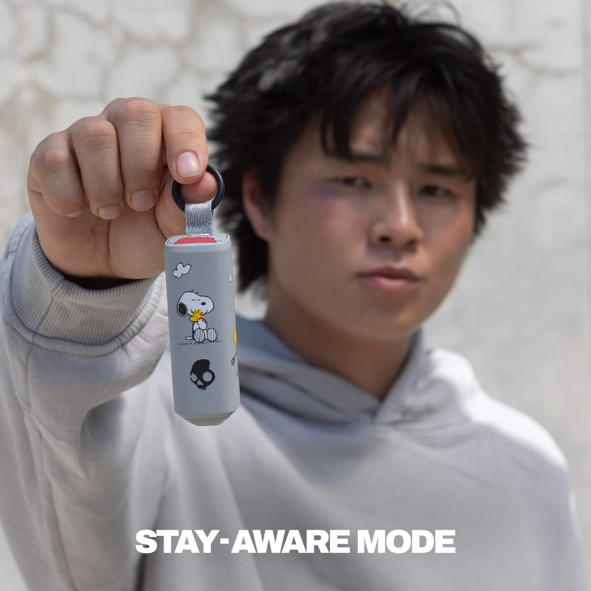 A person in a light gray hoodie holds up the dime 360 earbuds with woodstock-style cartoon graphics and a black loop at the top. The text 'STAY-AWARE MODE' appears at the bottom. | Skullcandy X Peanuts Woodstock & Snoopy