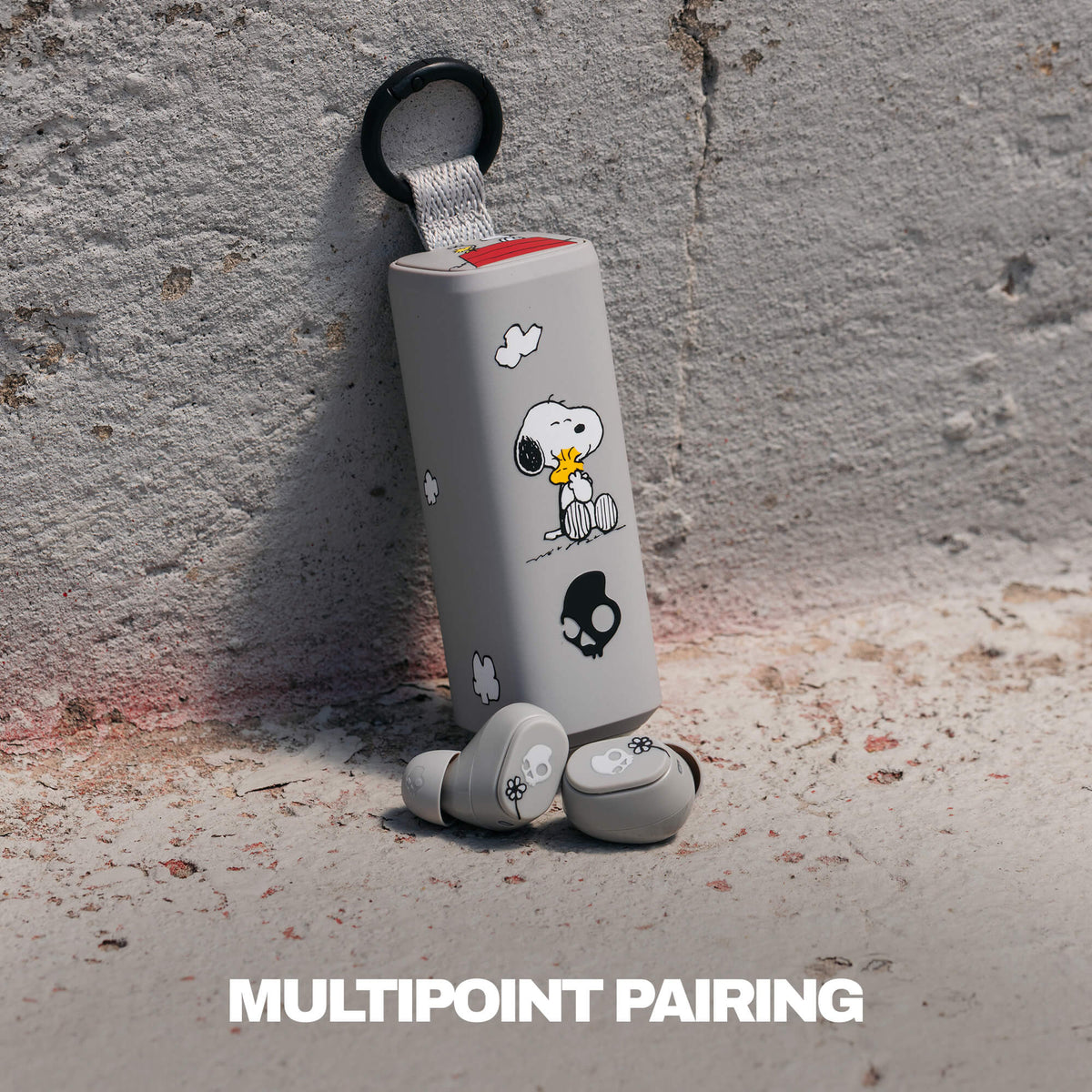 A pair of gray true wireless earbuds with a matching cylindrical charging case featuring woodstock illustrations, leaning against a textured concrete wall. The text 'MULTIPOINT PAIRING' is displayed at the bottom.  | Skullcandy X Peanuts Woodstock & Snoopy