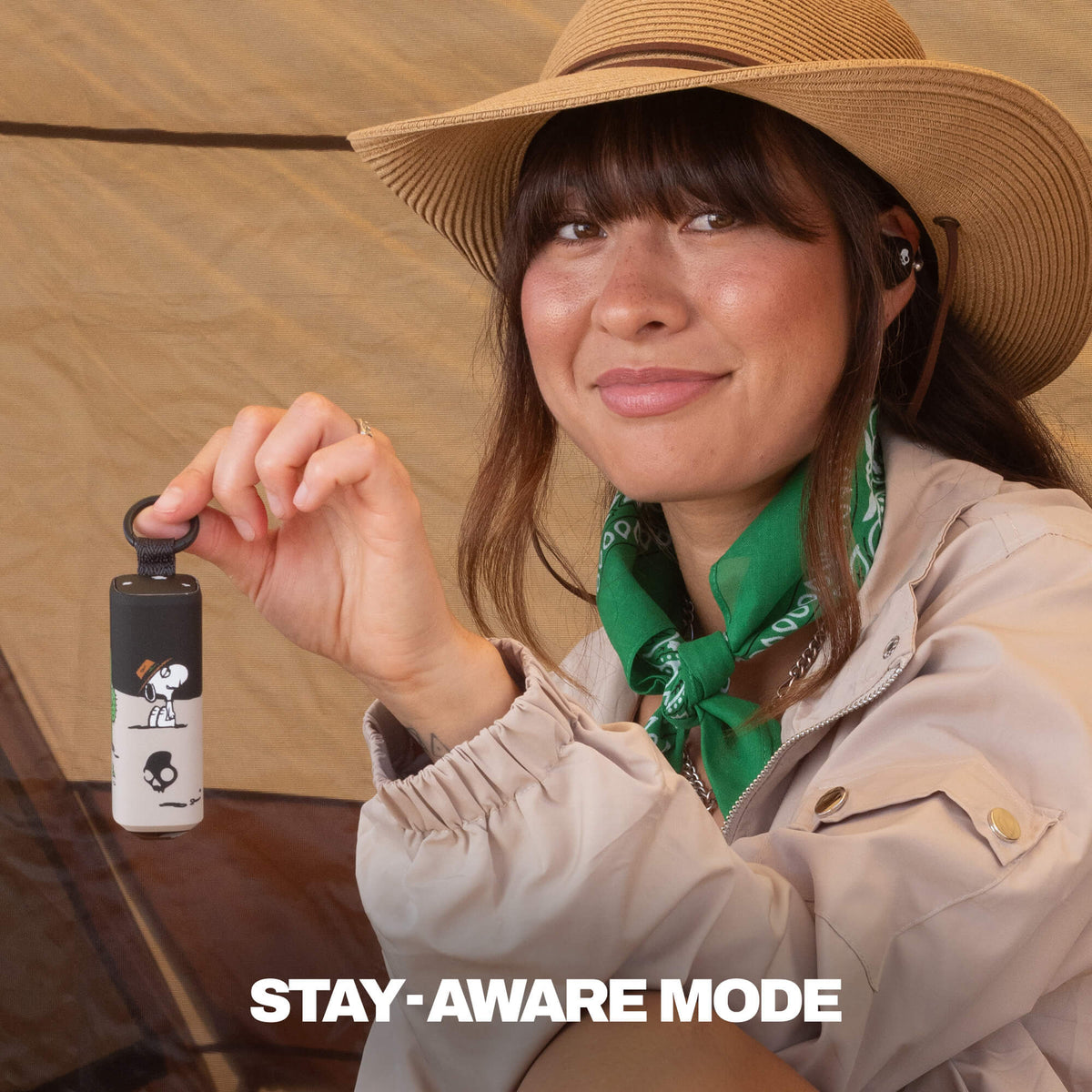 A woman wearing a tan wide-brim hat, beige jacket, and green bandana holds up a small black-and-white dime 360 earbud with a spike graphic and the text 'STAY-AWARE MODE' at the bottom.  | Skullcandy X Peanuts Spike
