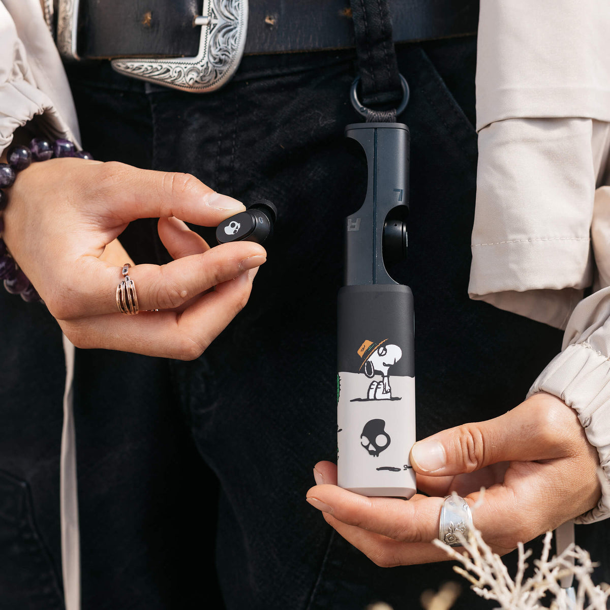 A close-up lifestyle shot of a person holding a black and white Dime 360 with spike and a small black earbud accessory, wearing a cream shirt and black pants.  | Skullcandy X Peanuts Spike