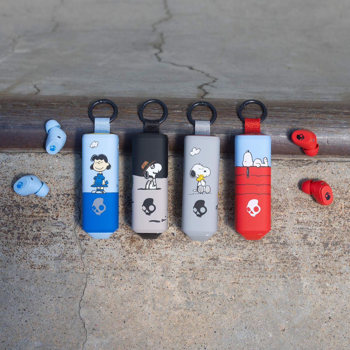 Four peanuts-themed keychain dime 360 earbuds in blue, black, gray, and red are lined up on a weathered wooden beam, with matching red and blue earbuds resting on either side on a concrete surface.  | Skullcandy X Peanuts Spike