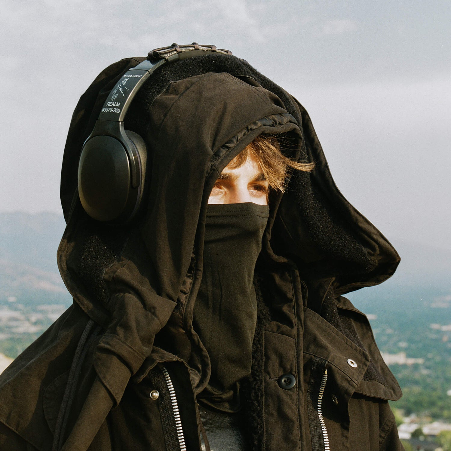 Person wearing a dark coat and headphones with a blurred landscape in the background | Dustbox