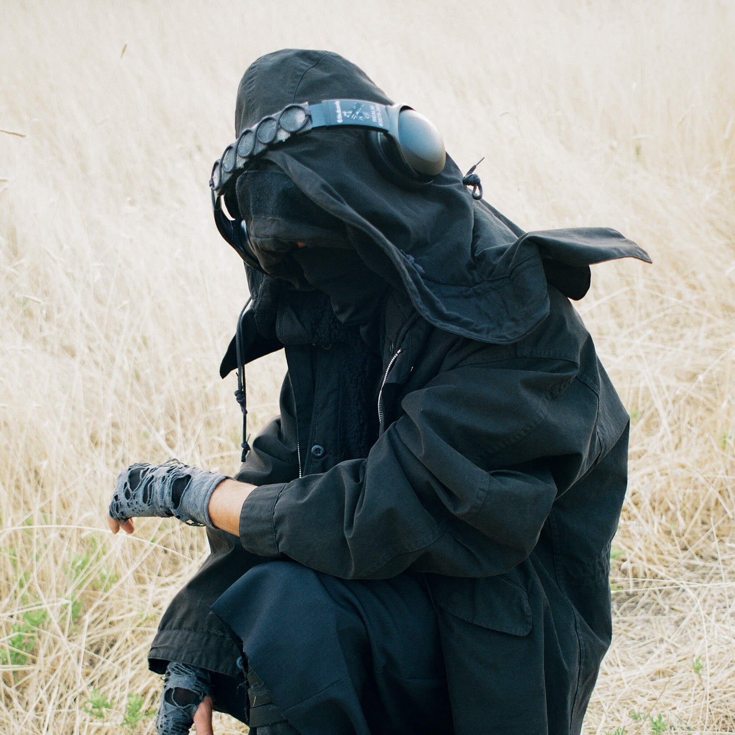 Person in black hooded outfit sitting in a field of dry grass | Dustbox