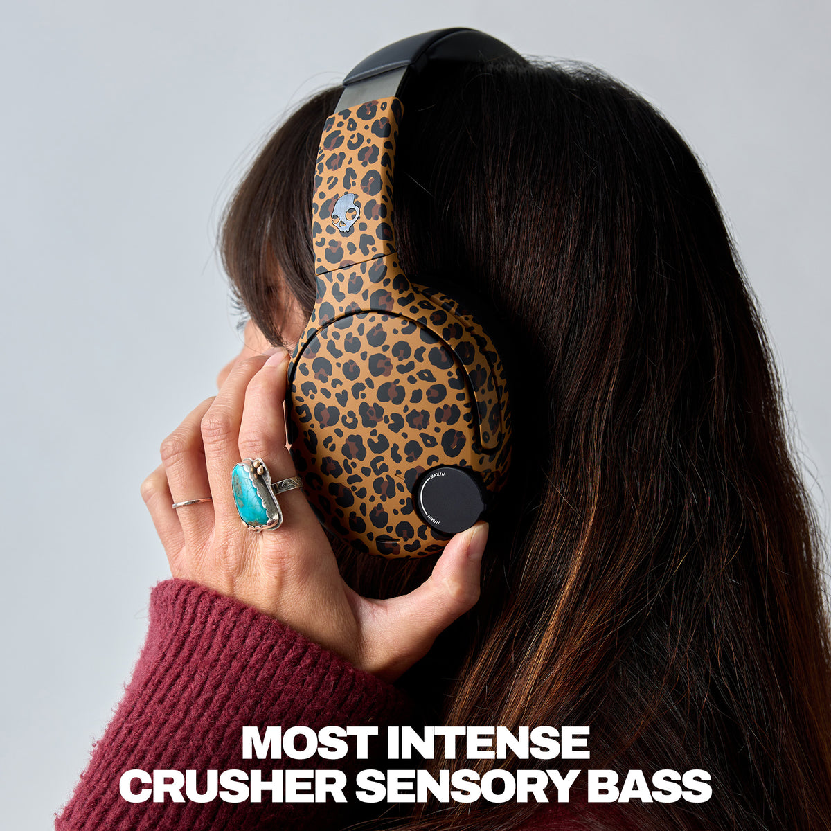 A person wearing leopard-print over-ear headphones with a dark burgundy knit sweater and a turquoise ring, shown from the side against a light gray background with the text 'MOST INTENSE CRUSHER SENSORY BASS' at the bottom. | Leopard