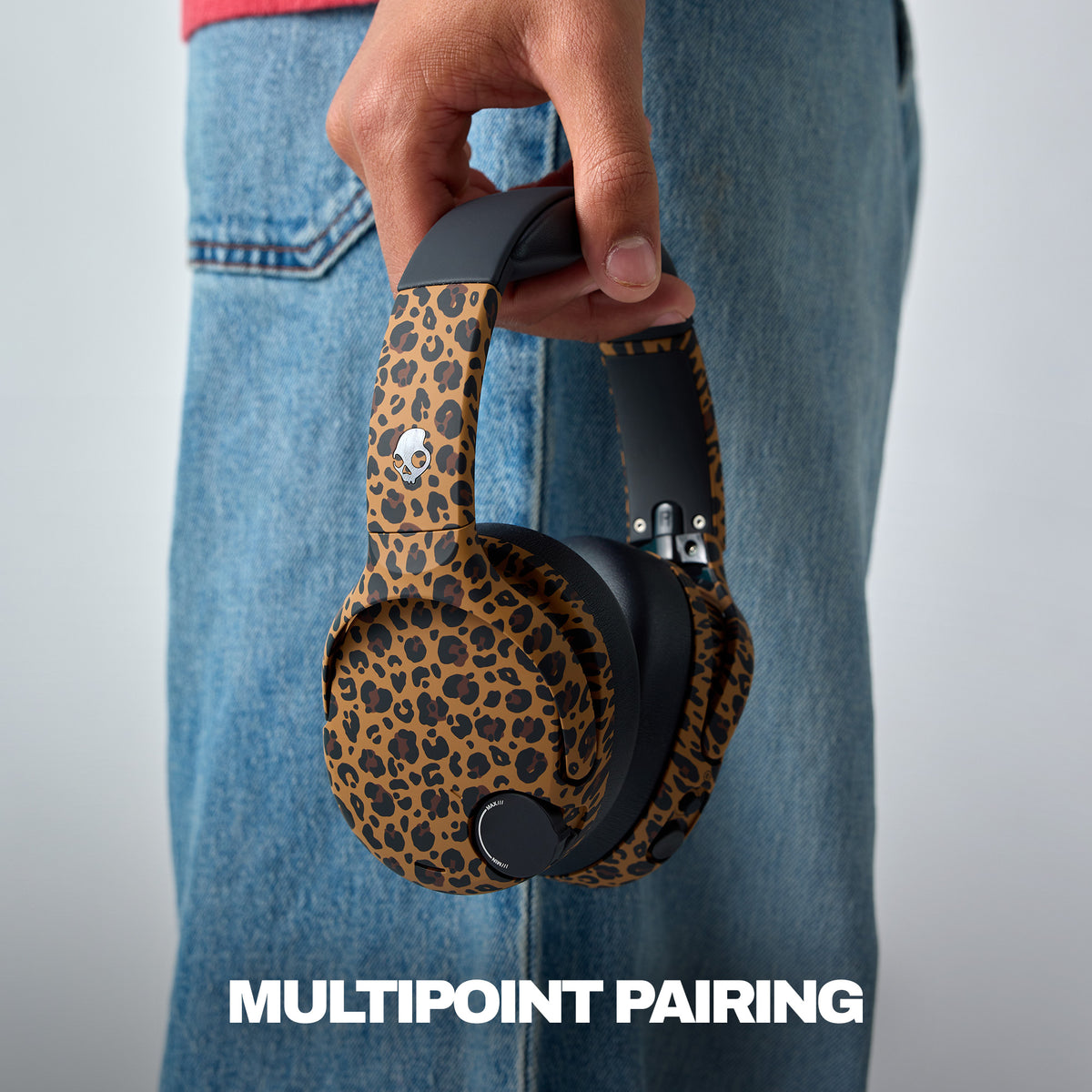 A person in blue jeans holds a pair of over-ear headphones with a brown and black leopard print fabric and black headband arms. The text 'MULTIPOINT PAIRING' is displayed at the bottom. | Leopard