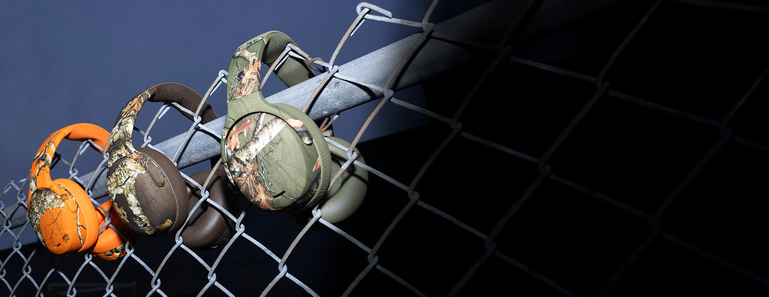 Three pairs of headphones in different colors hanging on a chain-link fence with a dark background.