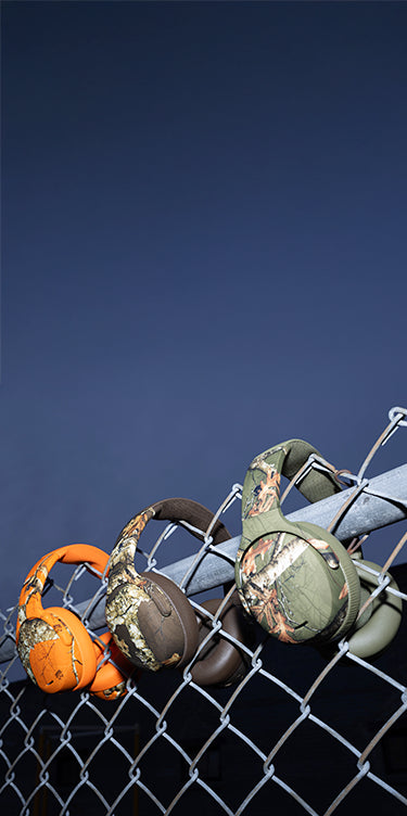 Three pairs of camouflage-patterned headphones on a chain-link fence against a blue sky.