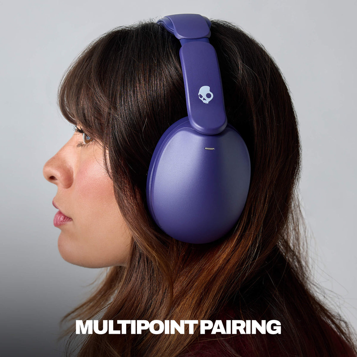 A woman in profile wearing a pair of hesh 360 over-ear headphones in a matte purple color, with the brand's skull logo visible on the headband. The image features a gray studio background and the text 'MULTIPOINT PAIRING' at the bottom.  | Future