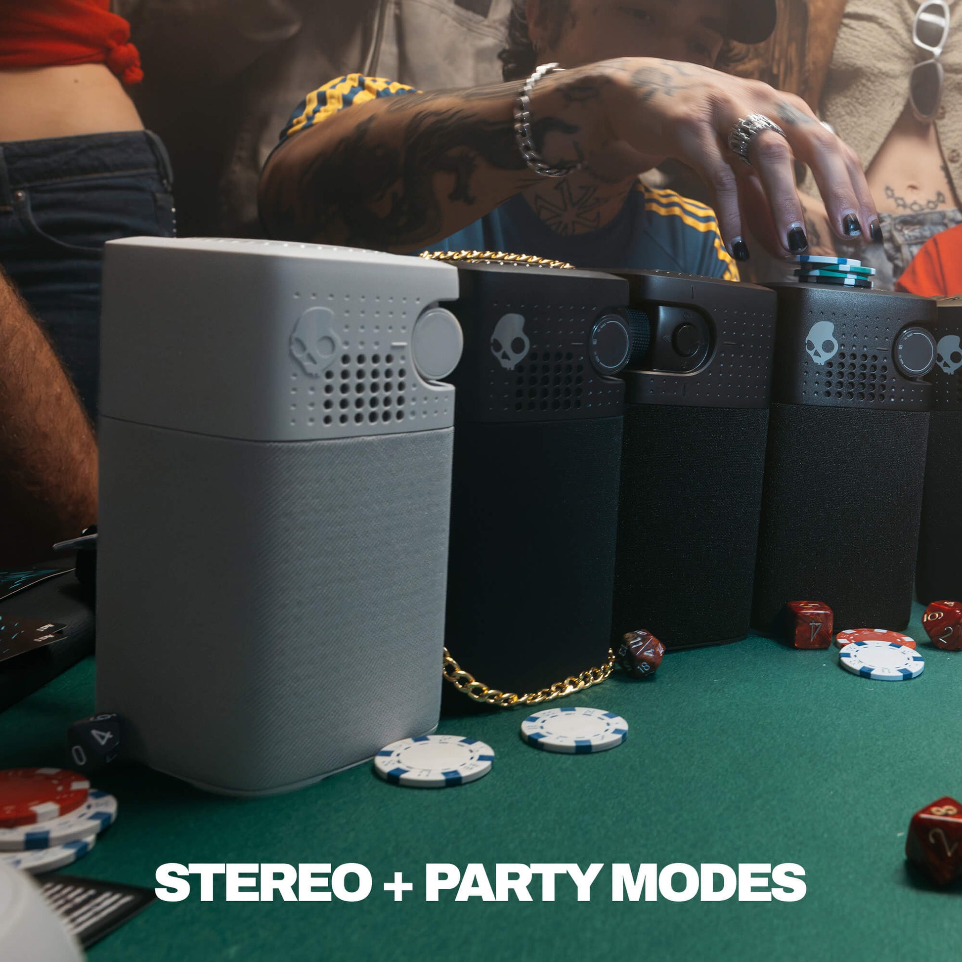 A group of session 900 speakers  arranged on a green felt table with poker chips and a person's tattooed hand reaching toward them. The devices are shown in gray and black, with one gold chain draped across them. | True Black