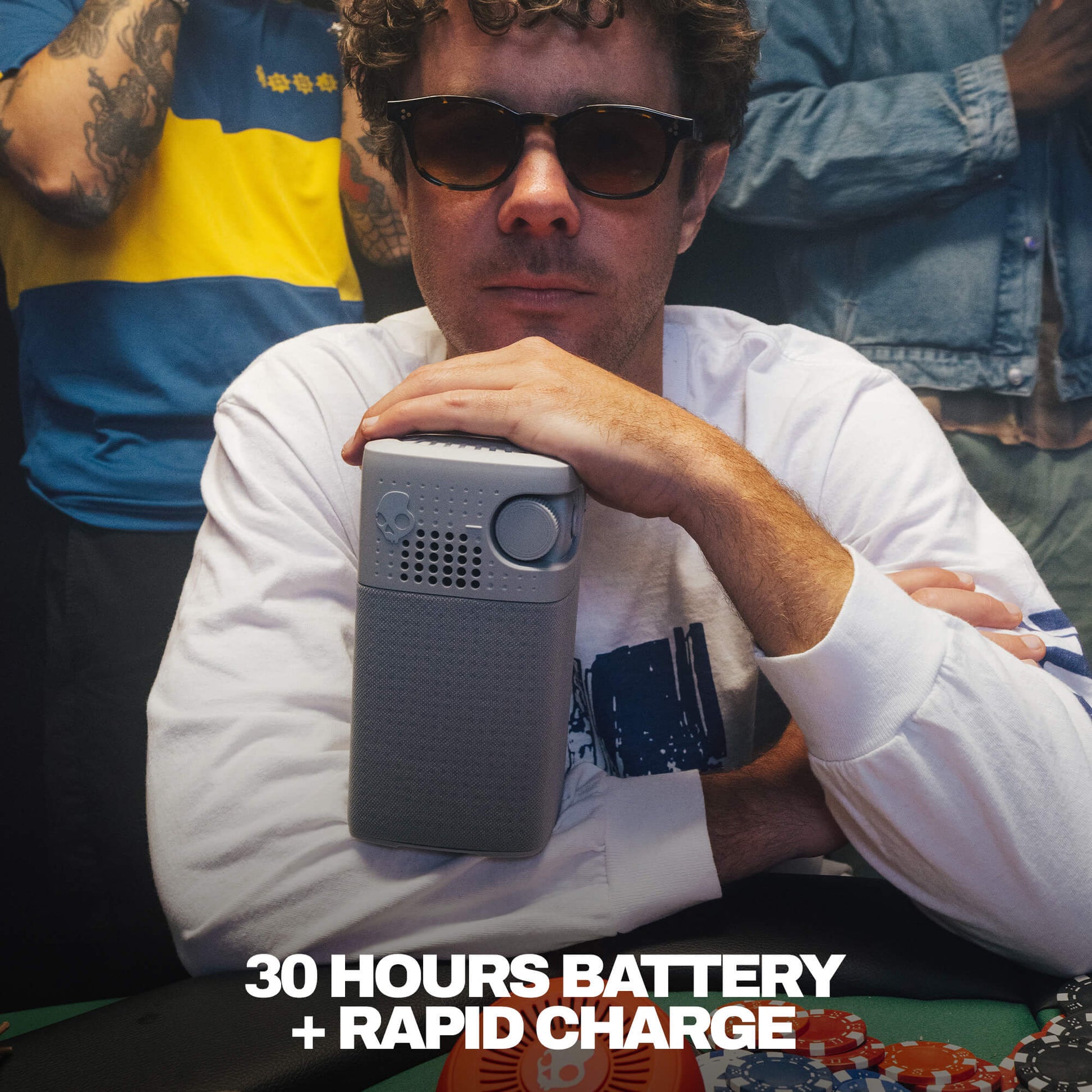 A man wearing sunglasses and a white shirt holds a gray session 900 speaker with a circular button, with text overlay reading '30 HOURS BATTERY + RAPID CHARGE'. | Cement