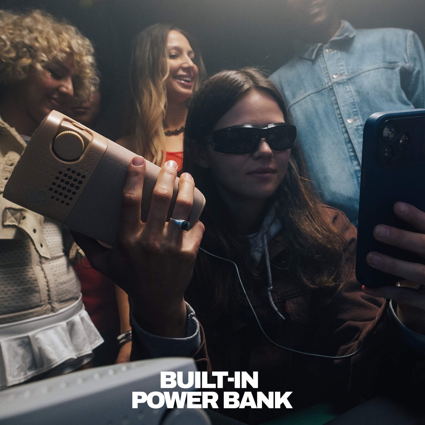 A woman wearing dark sunglasses holds a session 900 speaker in one hand while taking a selfie with a smartphone. Other people are visible in the background, and the text 'BUILT-IN POWER BANK' appears at the bottom. | Cement
