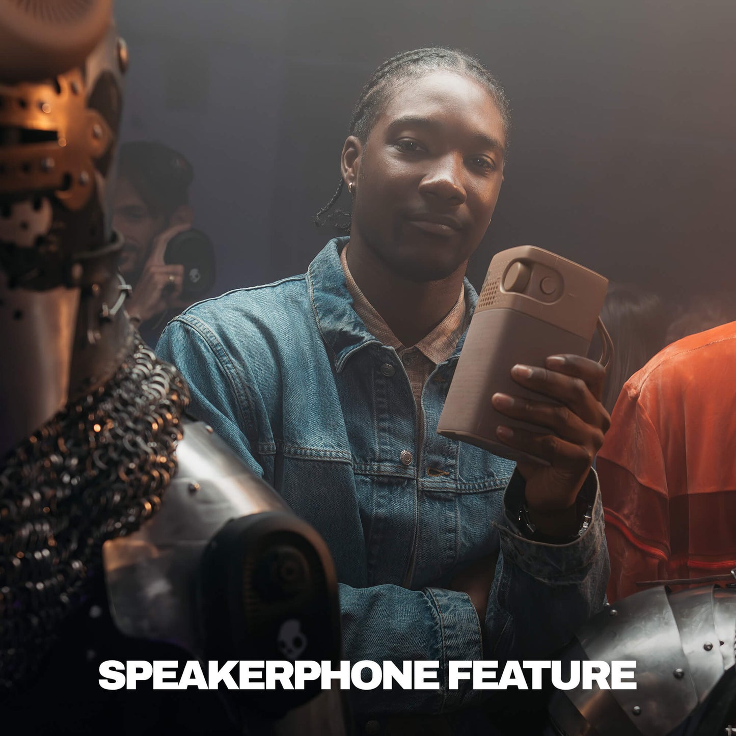 A person in a denim jacket holds a session 900 speaker in a dimly lit indoor setting, with armored figures partially visible on both sides. White text at the bottom reads 'SPEAKERPHONE FEATURE'. | Primer