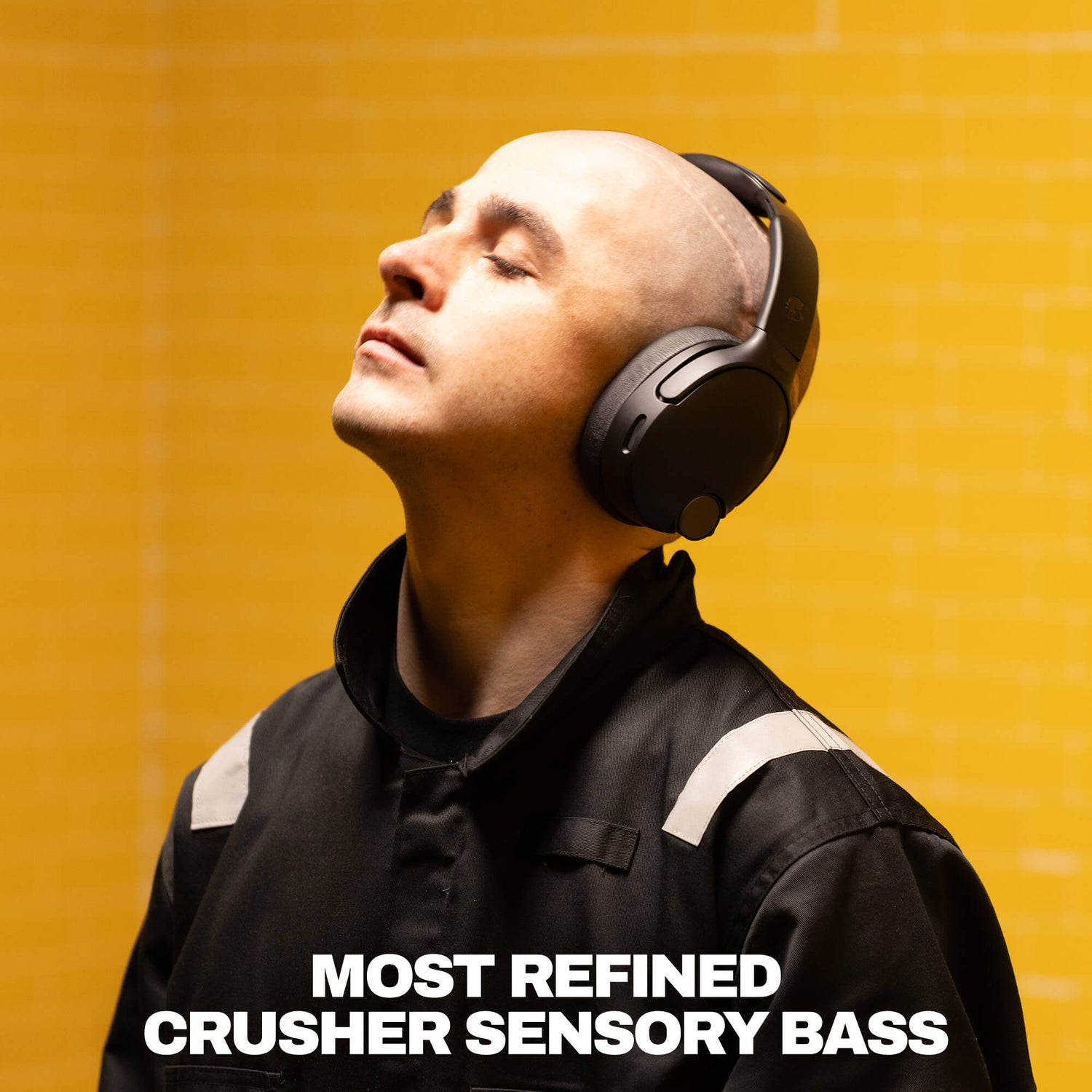 A man wearing black over-ear headphones and a black jacket with white shoulder stripes, looking upward against a bright yellow background. Large white text at the bottom reads 'MOST REFINED CRUSHER SENSORY BASS'. | True Black