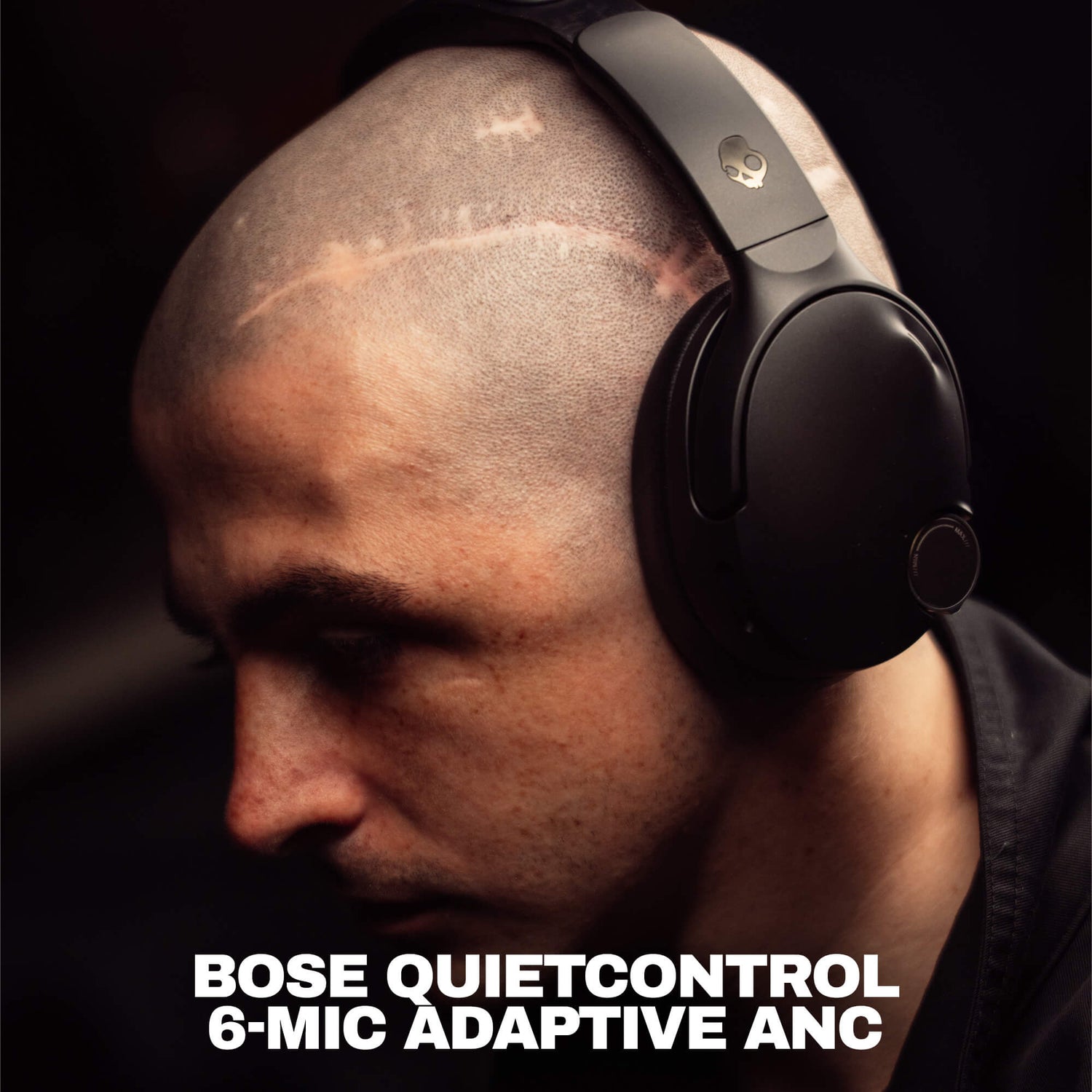 A close-up of a man wearing black Crusher 1080 headphones with Bose QuietControl 6-Mic Adaptive ANC over-ear headphones against a dark background, with bold white text at the bottom reading 'BOSE QUIETCONTROL 6-MIC ADAPTIVE ANC'. | True Black