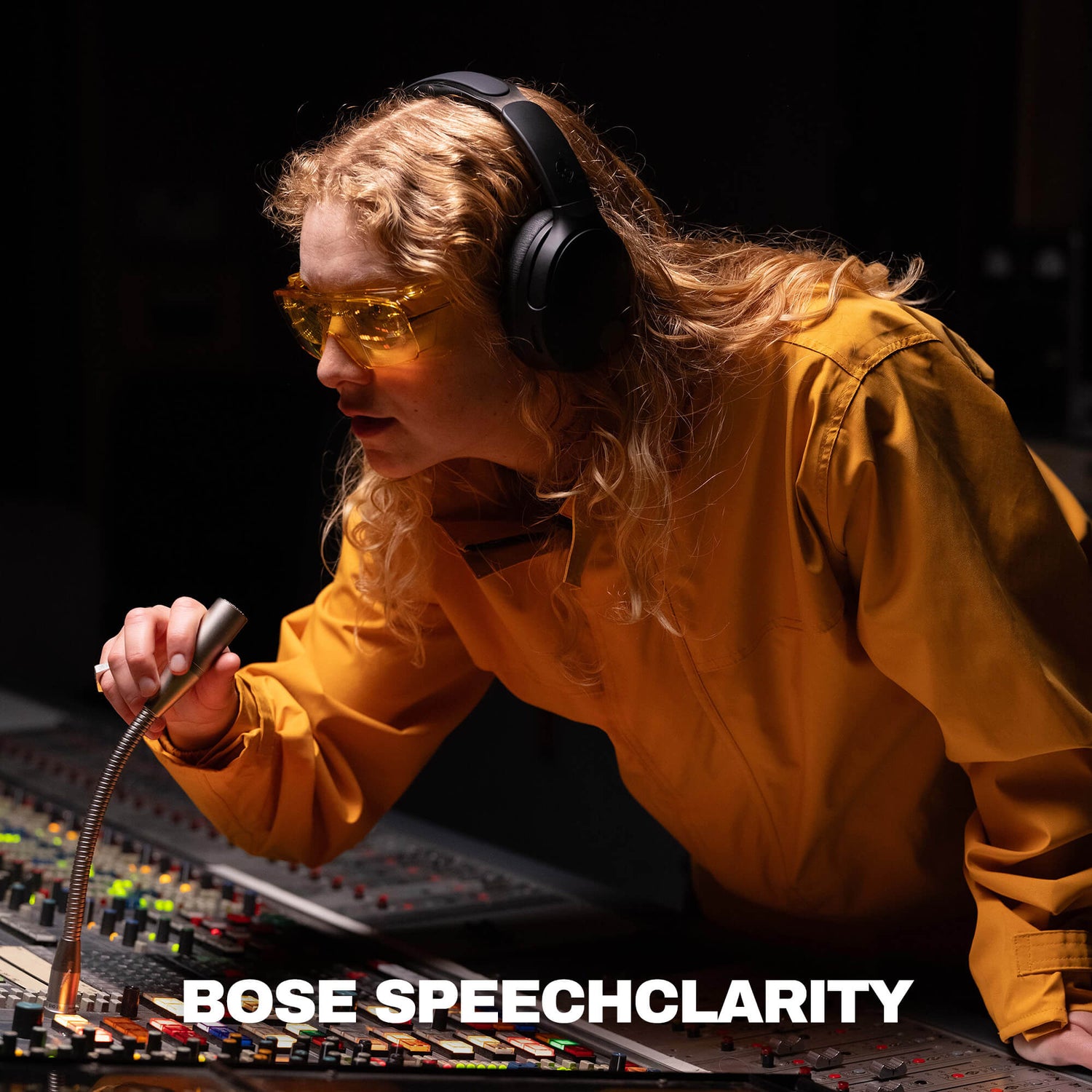 A woman in a recording studio wearing yellow-tinted safety glasses and black Crusher 1080 over-ear headphones, leaning over a mixing board and holding a microphone. The text 'BOSE SPEECHCLARITY' appears at the bottom. | True Black