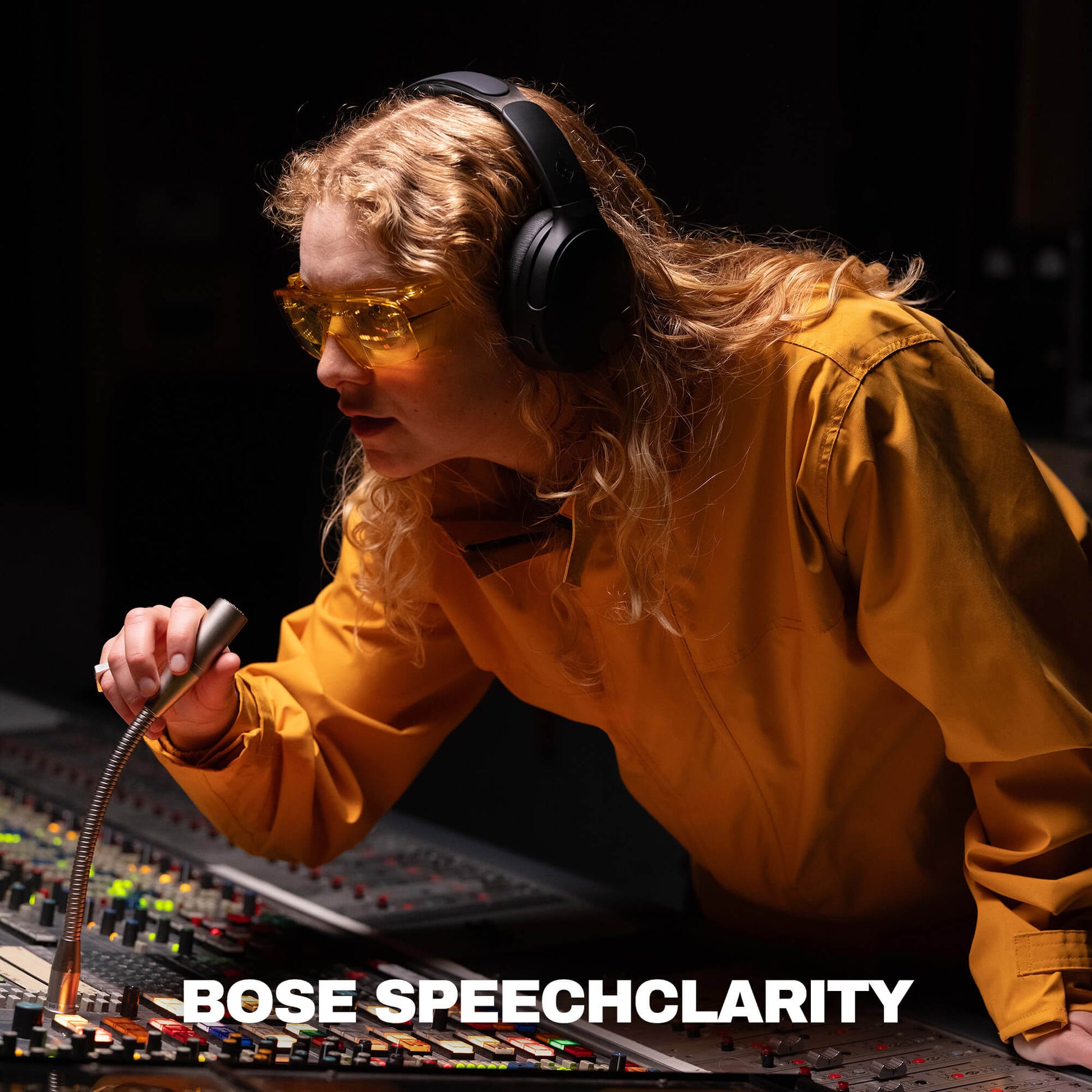 A woman in a recording studio wearing yellow-tinted safety glasses and black Crusher 1080 over-ear headphones, leaning over a mixing board and holding a microphone. The text 'BOSE SPEECHCLARITY' appears at the bottom. | True Black