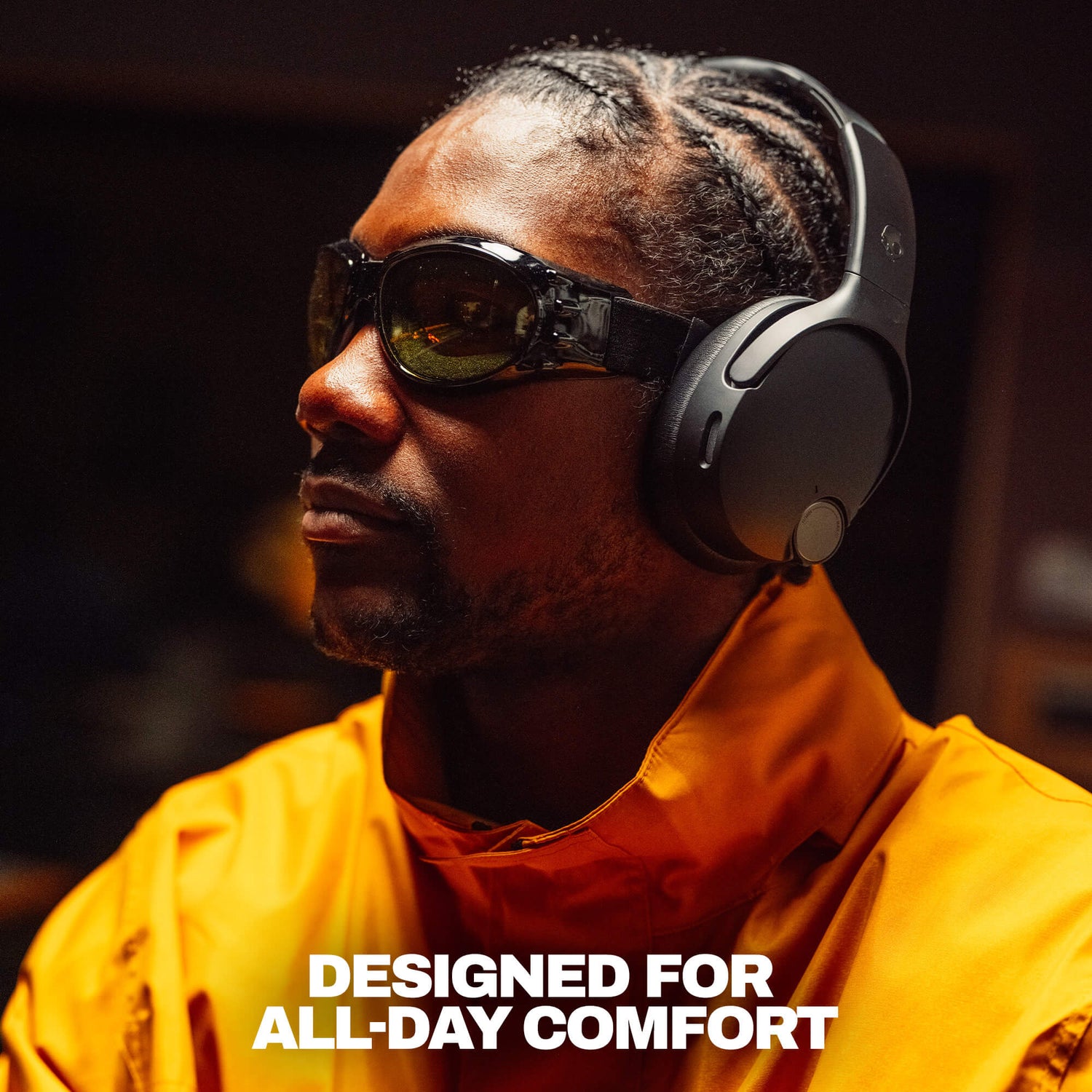 A man in a bright orange jacket wearing black over-ear headphones and dark sunglasses, shown in a dim indoor setting with the text 'DESIGNED FOR ALL-DAY COMFORT' at the  bottom. | True Black
