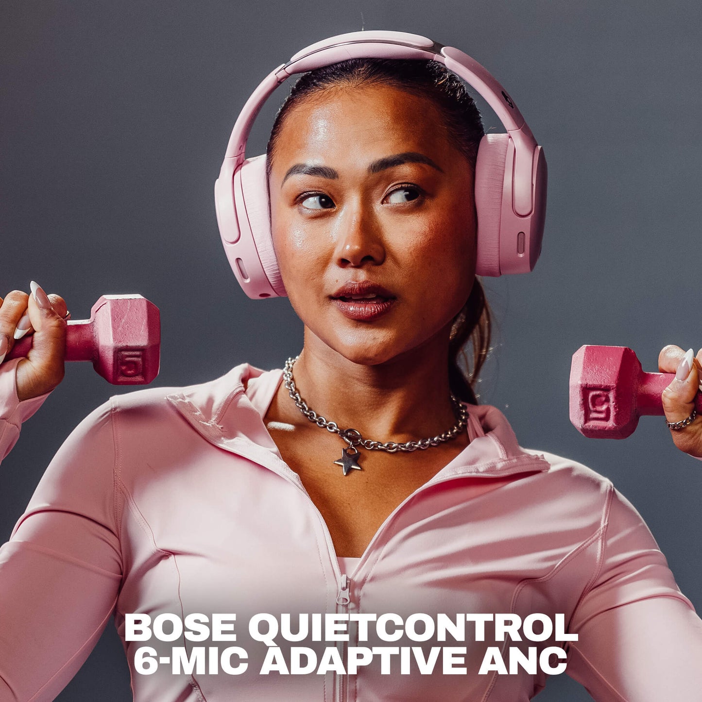 A woman wearing pink Crusher 1080 headphones with Bose QuietControl 6-MIC Adaptive ANC over-ear headphones and a matching pink athletic zip-up top, holding pink dumbbells on both sides against a dark gray studio background. White text at the bottom reads 'BOSE QUIETCONTROL 6-MIC ADAPTIVE ANC'. | Candy
