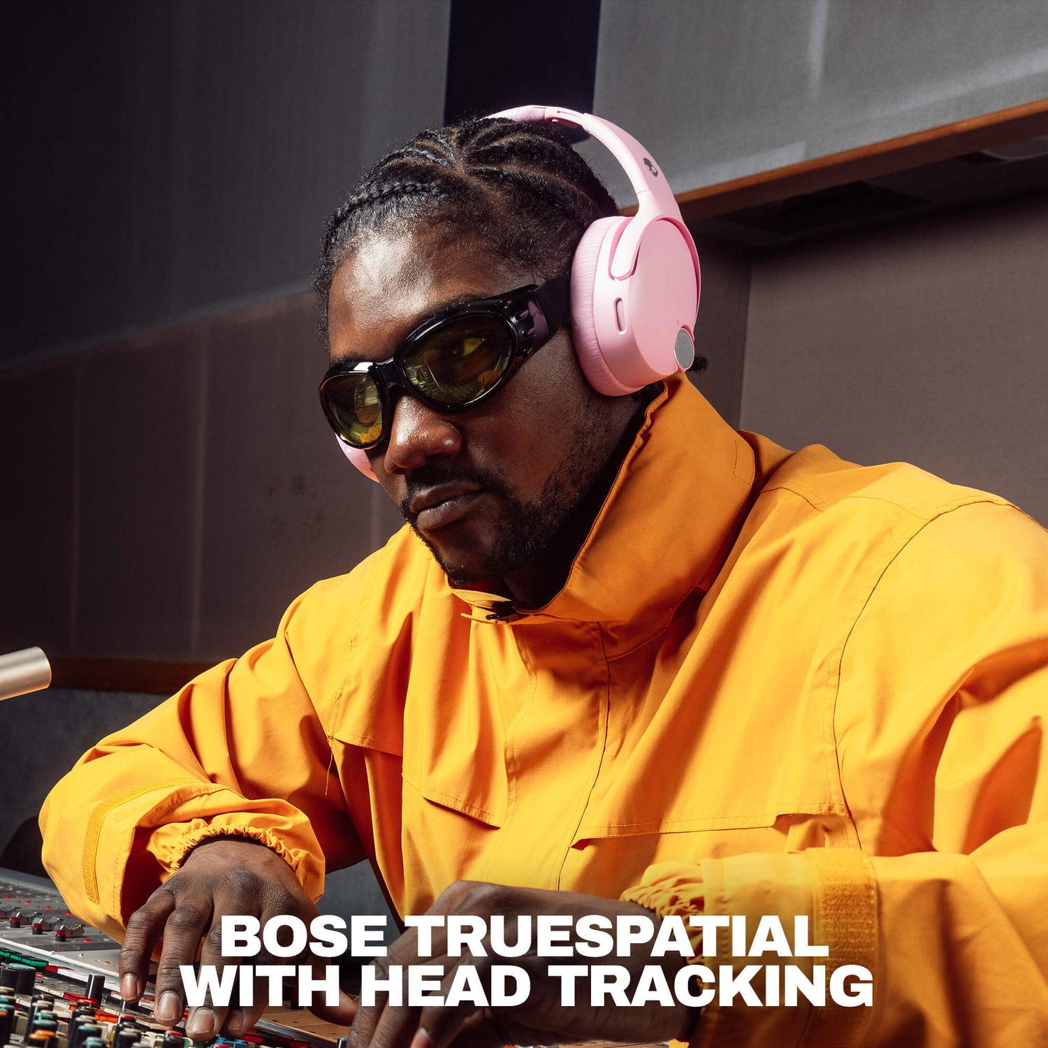 A man in a recording studio wearing pinkSkullcandy Crusher 1080 headphones with  Bose TrueSpatial headphones with head tracking, a bright yellow jacket, and dark sunglasses, with the text 'BOSE TRUESPATIAL WITH HEAD TRACKING' at the bottom. | Candy