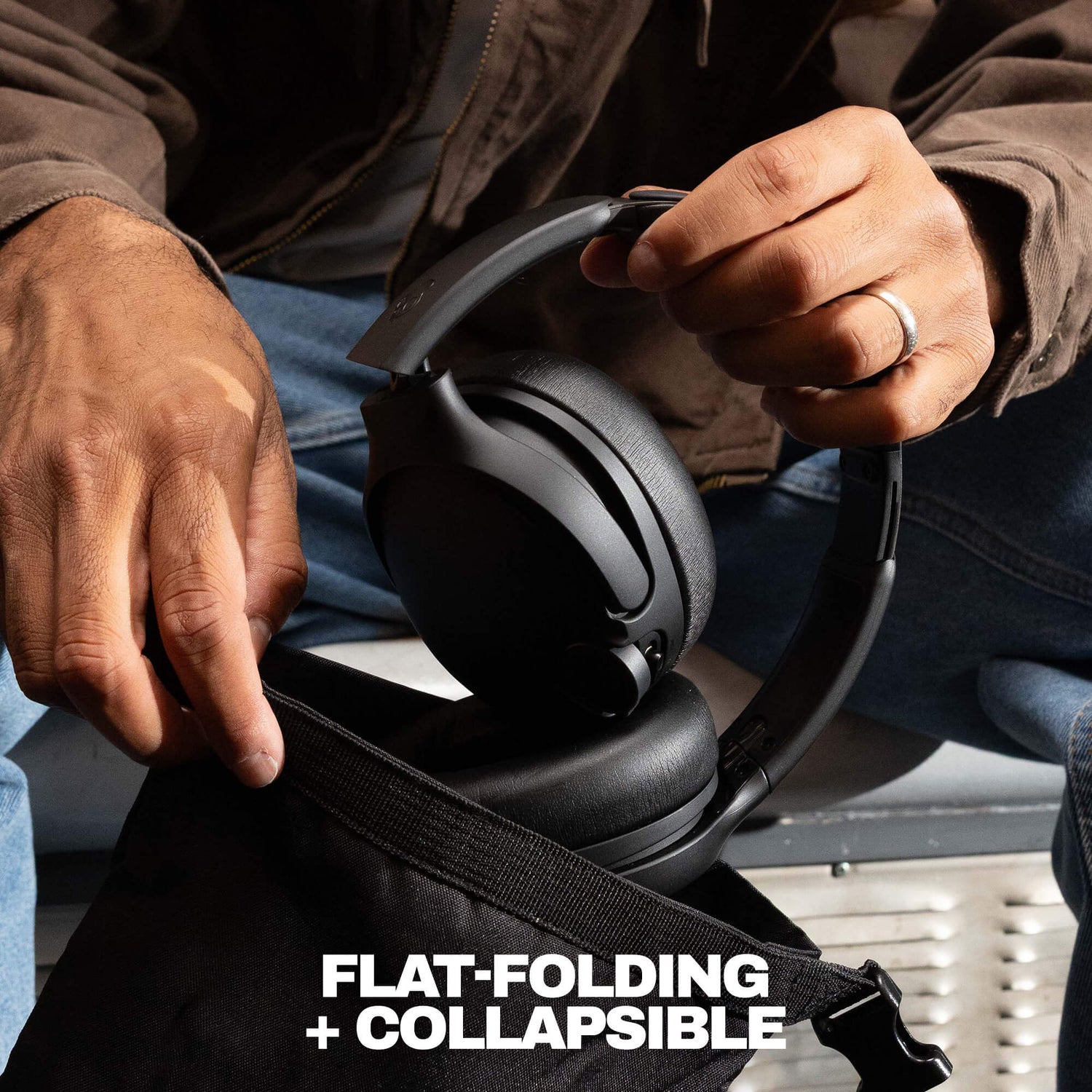 A close-up of a person holding a black over-ear headset over an open black bag, with the text 'FLAT-FOLDING + COLLAPSIBLE' at the bottom. The person is wearing a brown jacket and blue jeans. | Candy