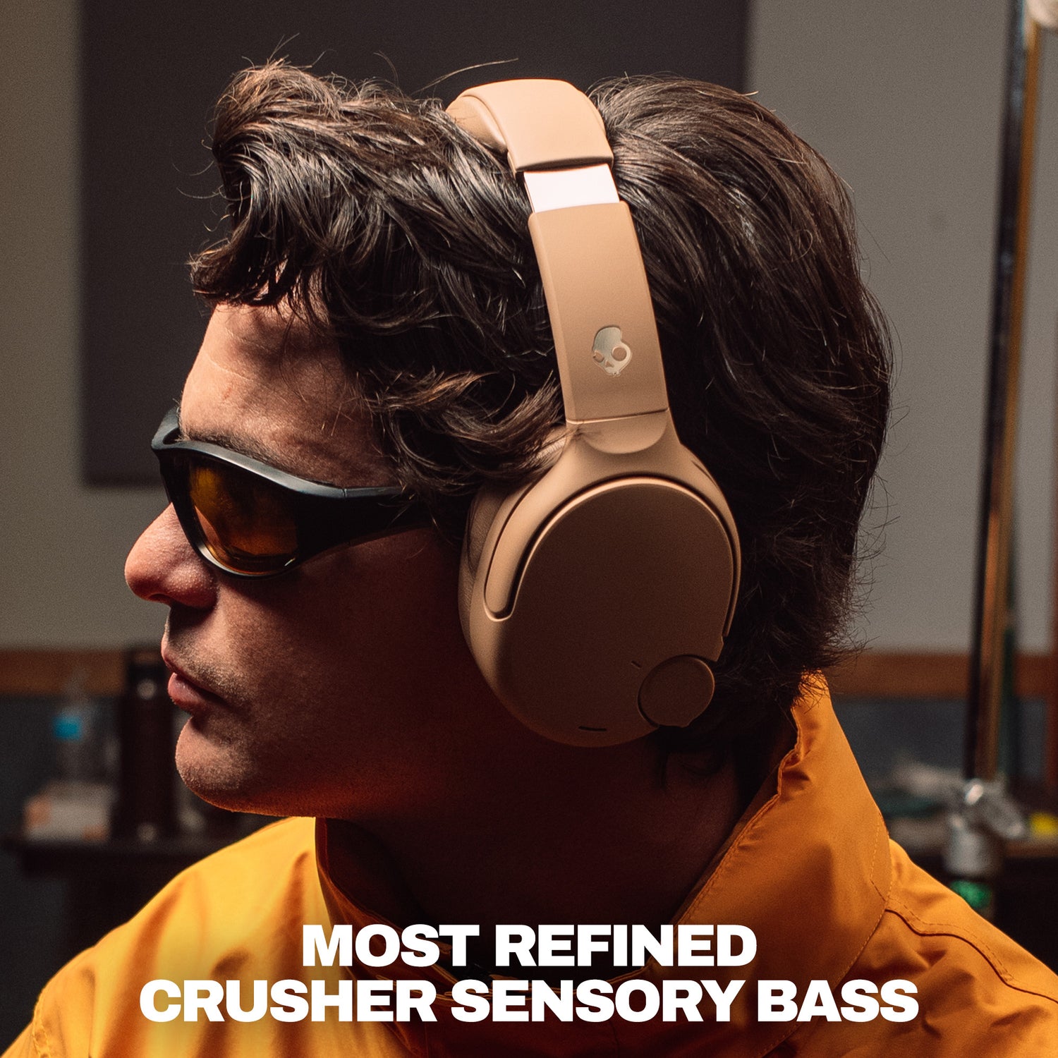 A man in profile wearing tan crusher 1080 over-ear headphones and dark sunglasses, with bold text at the bottom reading 'MOST REFINED CRUSHER SENSORY BASS'. | Primer