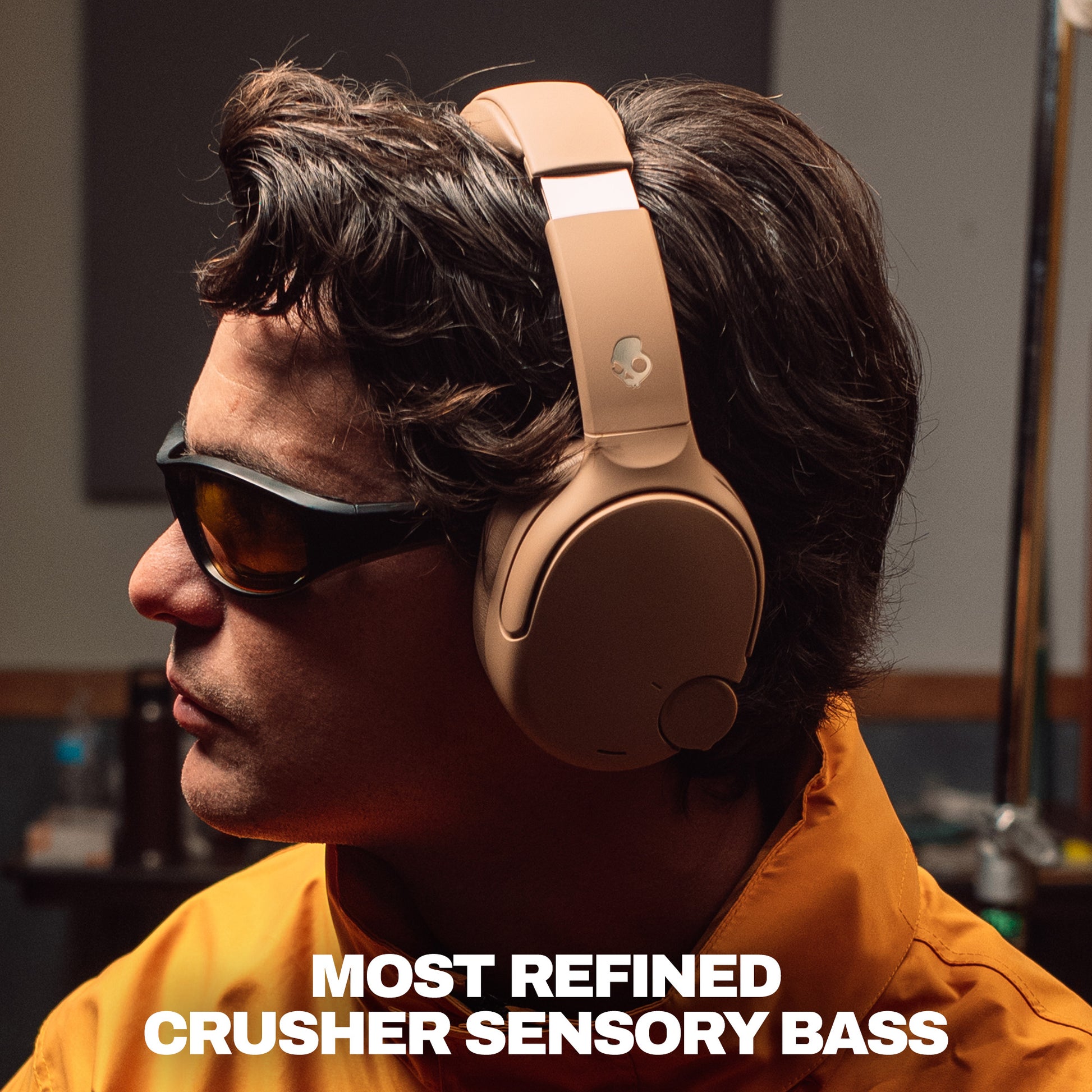 A man in profile wearing tan crusher 1080 over-ear headphones and dark sunglasses, with bold text at the bottom reading 'MOST REFINED CRUSHER SENSORY BASS'. | Primer