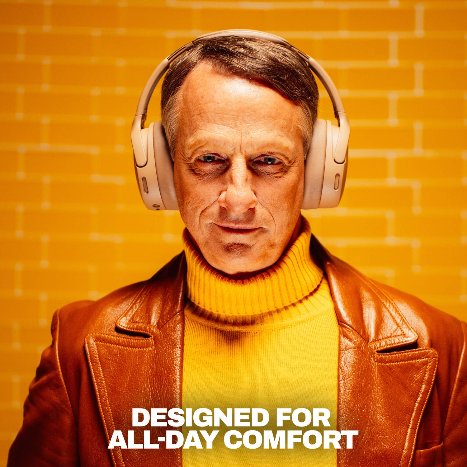 tony hawk wearing beige crusher 1080 over-ear headphones, a brown leather jacket, and a yellow turtleneck sweater stands against a yellow brick wall background with the text 'DESIGNED FOR ALL-DAY COMFORT' at the bottom. | Primer