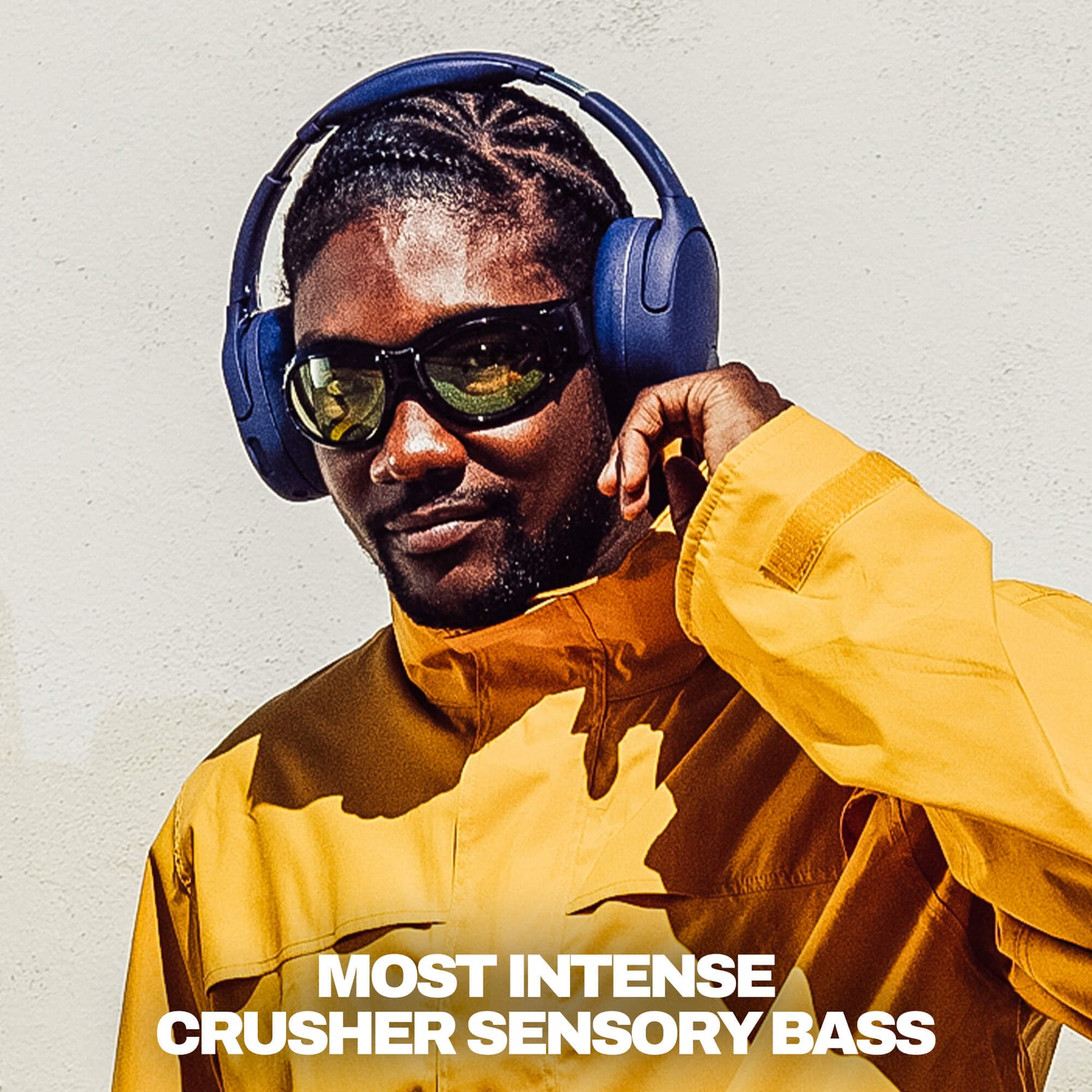 A man wearing deep blue Crusher 720 over-ear headphones,  sunglasses, dressed in a mustard yellow jacket, against a white wall. White text at the bottom reads 'MOST INTENSE CRUSHER SENSORY BASS'. | Future