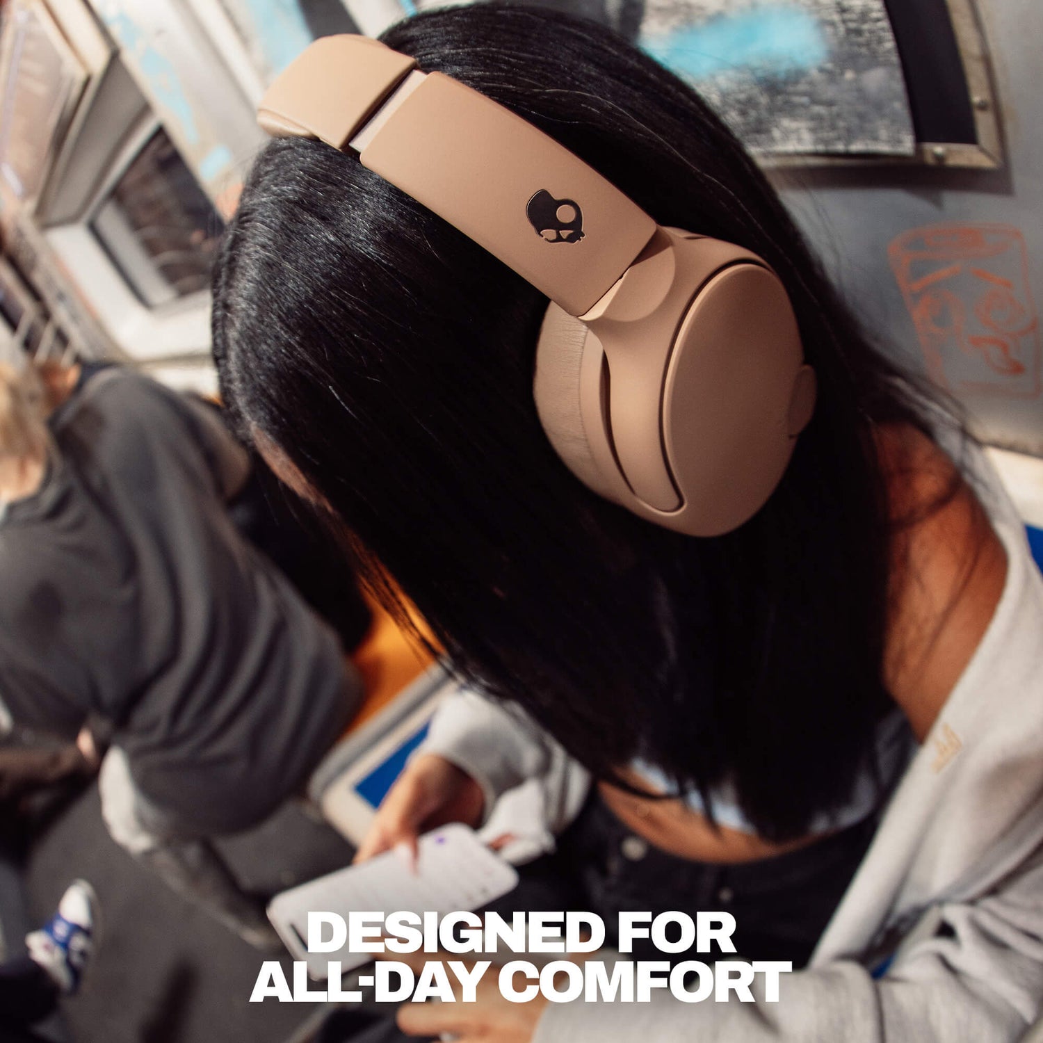 A person with long black hair wearing beige over-ear headphones on a subway train, with text 'DESIGNED FOR ALL-DAY COMFORT'. | Primer